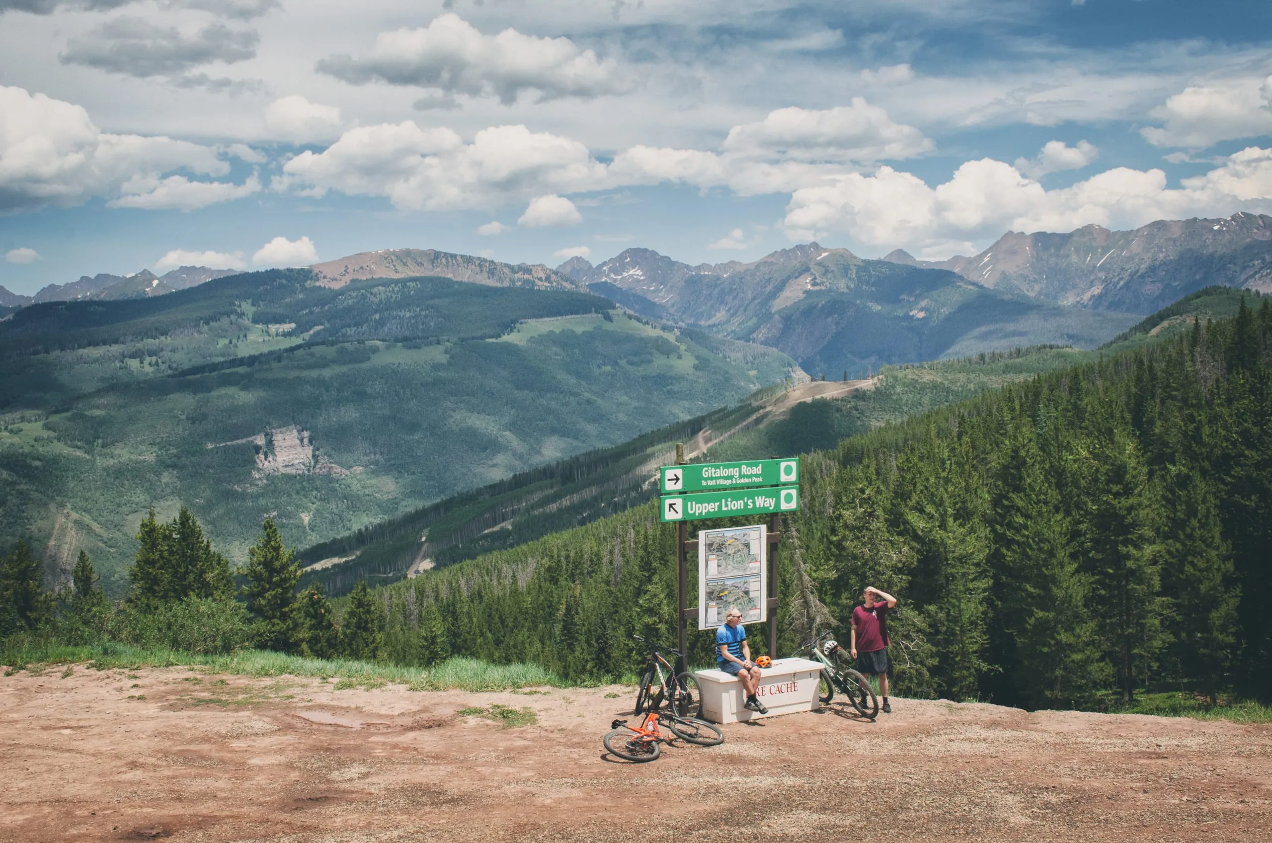 Two mountain bikers rest at a trail junction sign above a sweeping green Colorado mountain panorama.