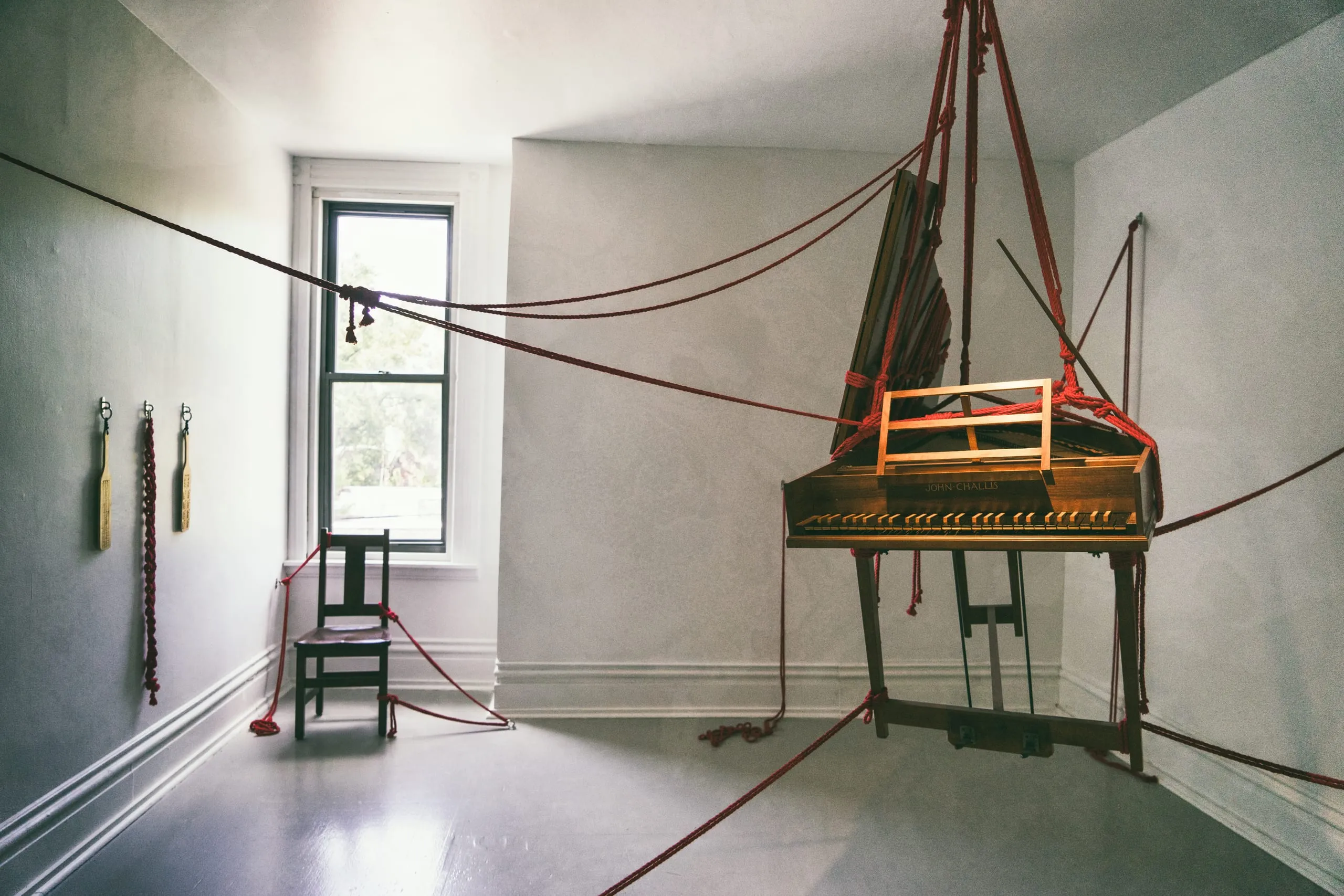 A wooden harpsichord hangs from a web of red ropes in a bare gray gallery room in Pittsburgh.