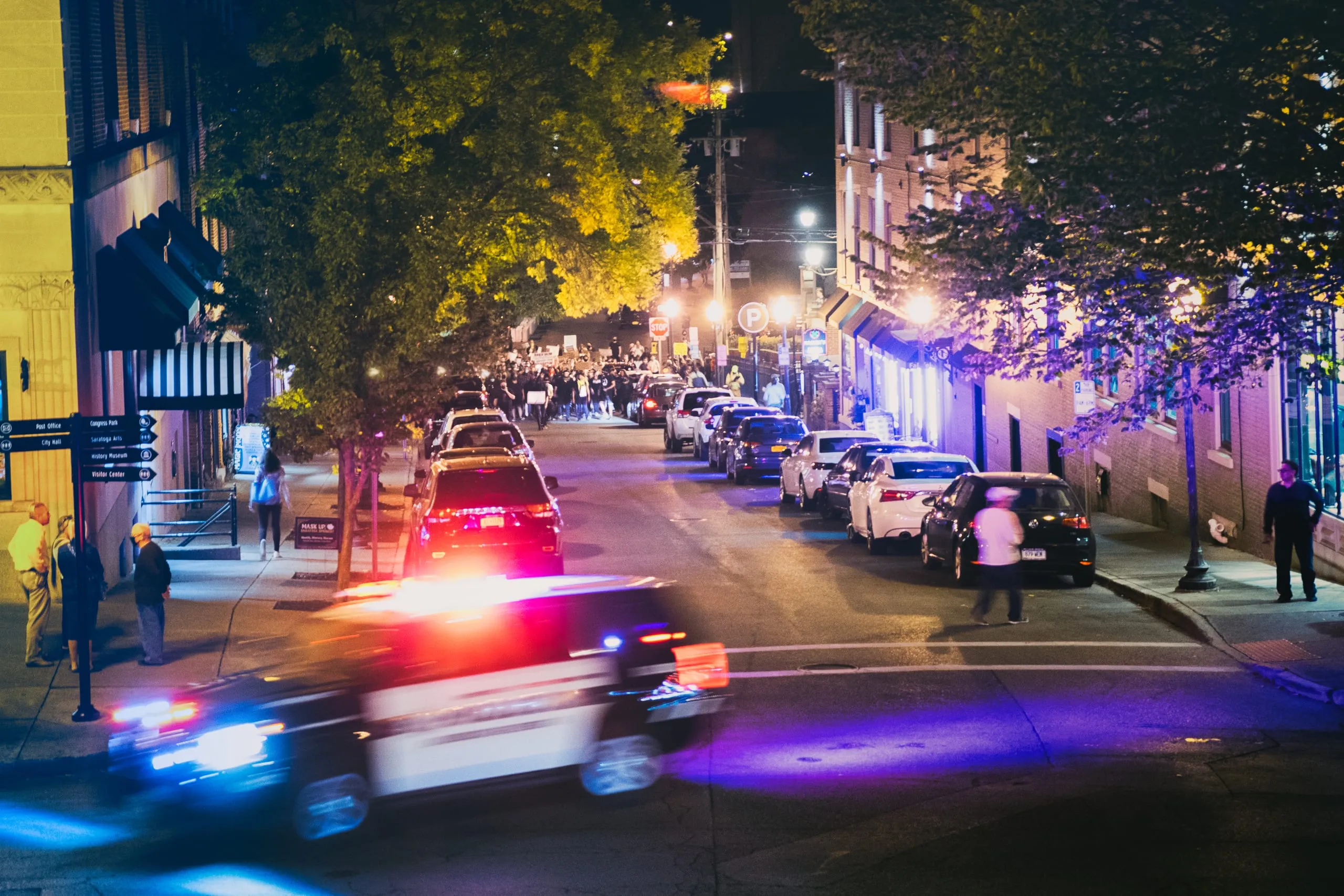 A police car with flashing lights blurs down a Saratoga Springs street at night as a crowd marches behind it.