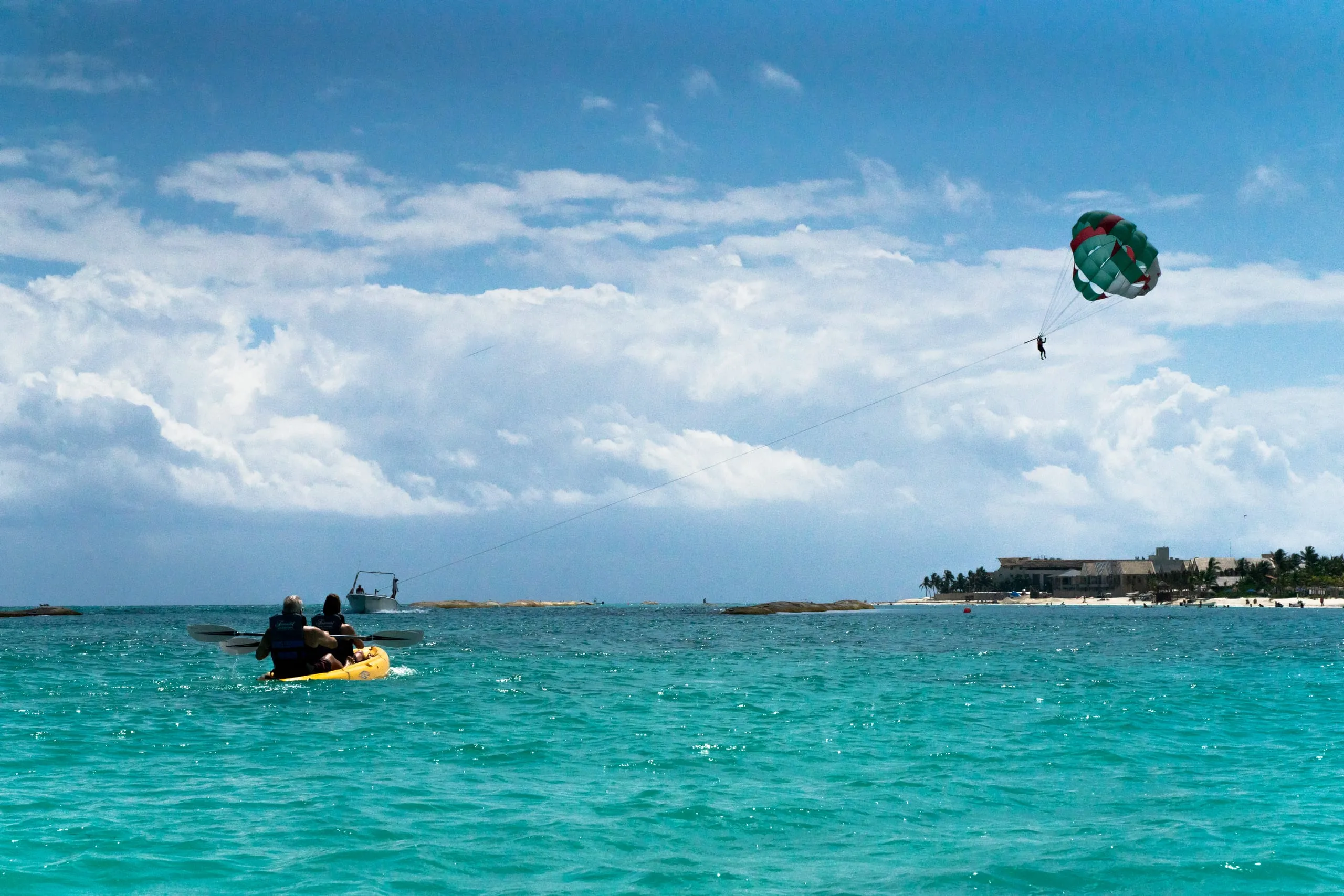 Two kayakers paddle turquoise water as a parasailer trails a boat toward a distant beach in Playa del Carmen.