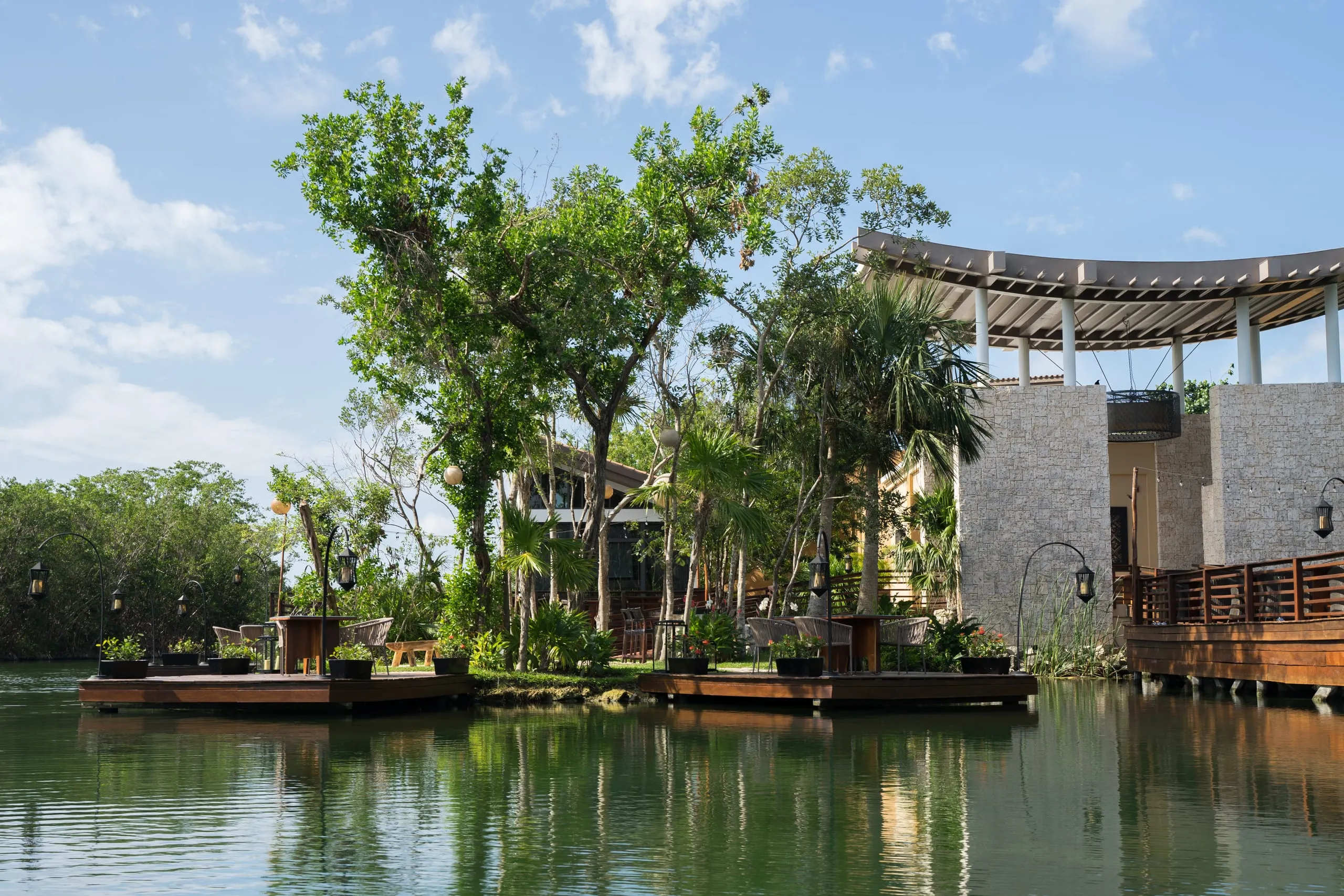 Wooden decks and a curved stone pavilion sit among trees along a green lagoon in Playa del Carmen.