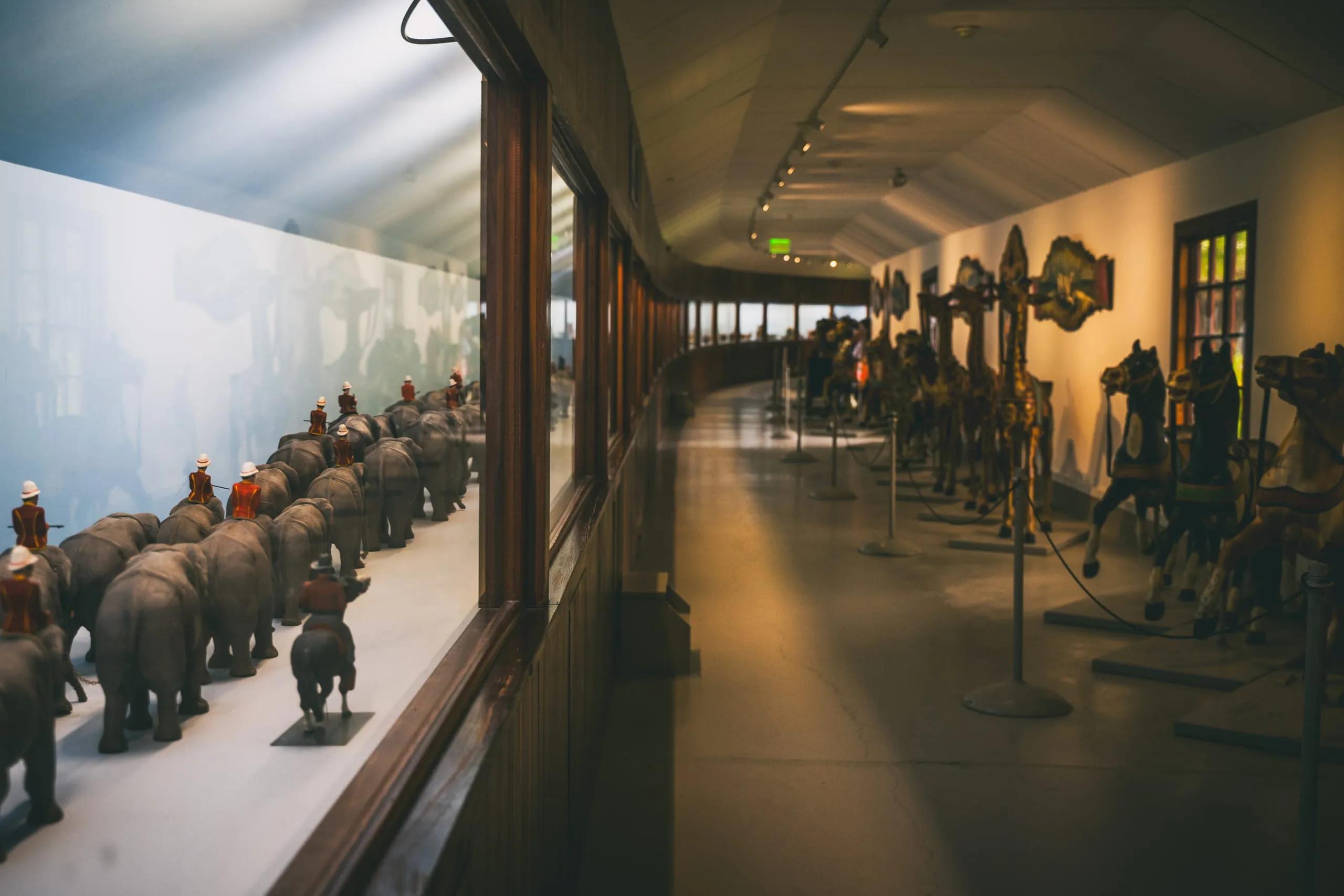 A curving museum gallery in Vermont with a diorama of tiny carved circus elephants behind glass and carousel horses on stands opposite.