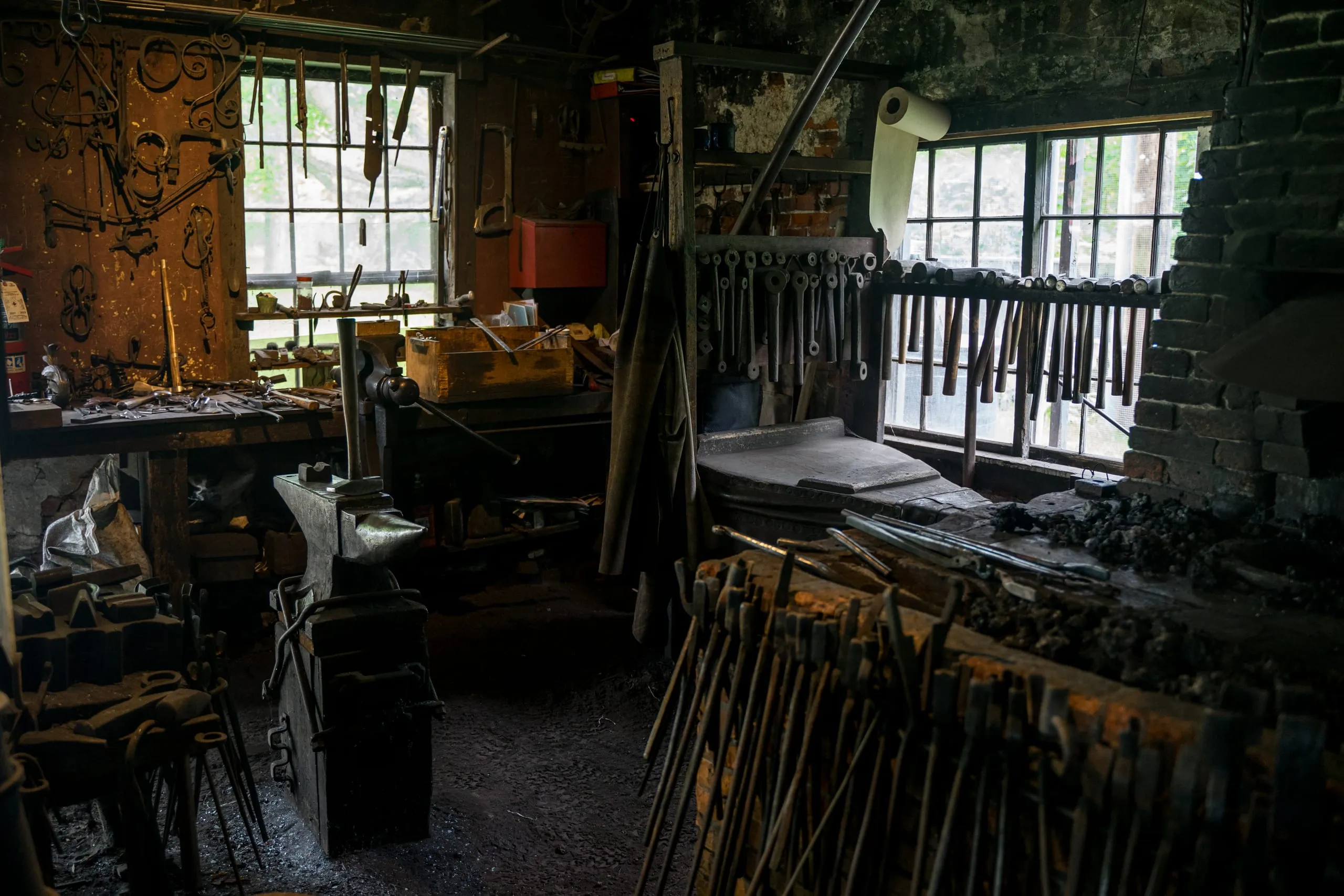Dim blacksmith workshop in Vermont with an anvil, vises, and hand tools hung by the windows.