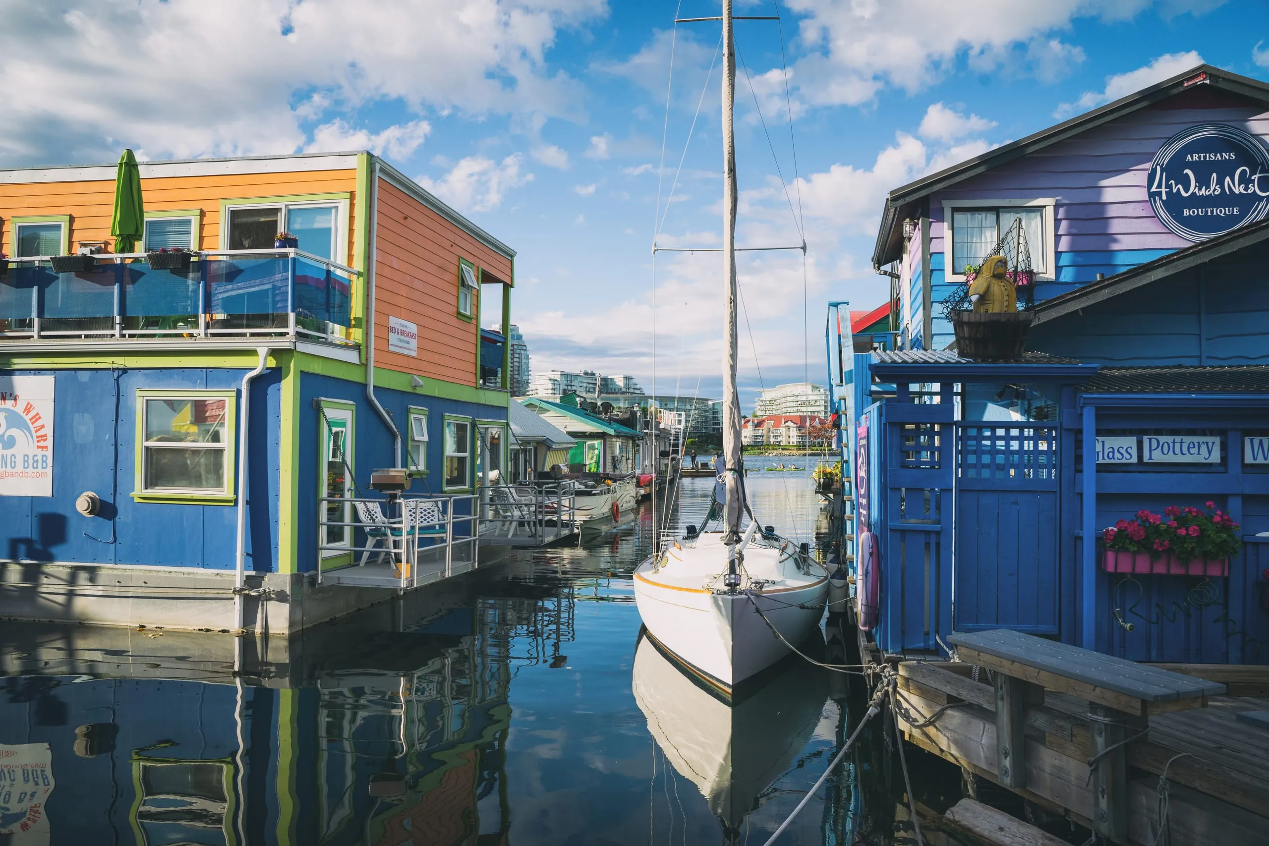 A white sailboat moored between brightly painted floating homes and shops on the water at Fisherman's Wharf in Victoria.