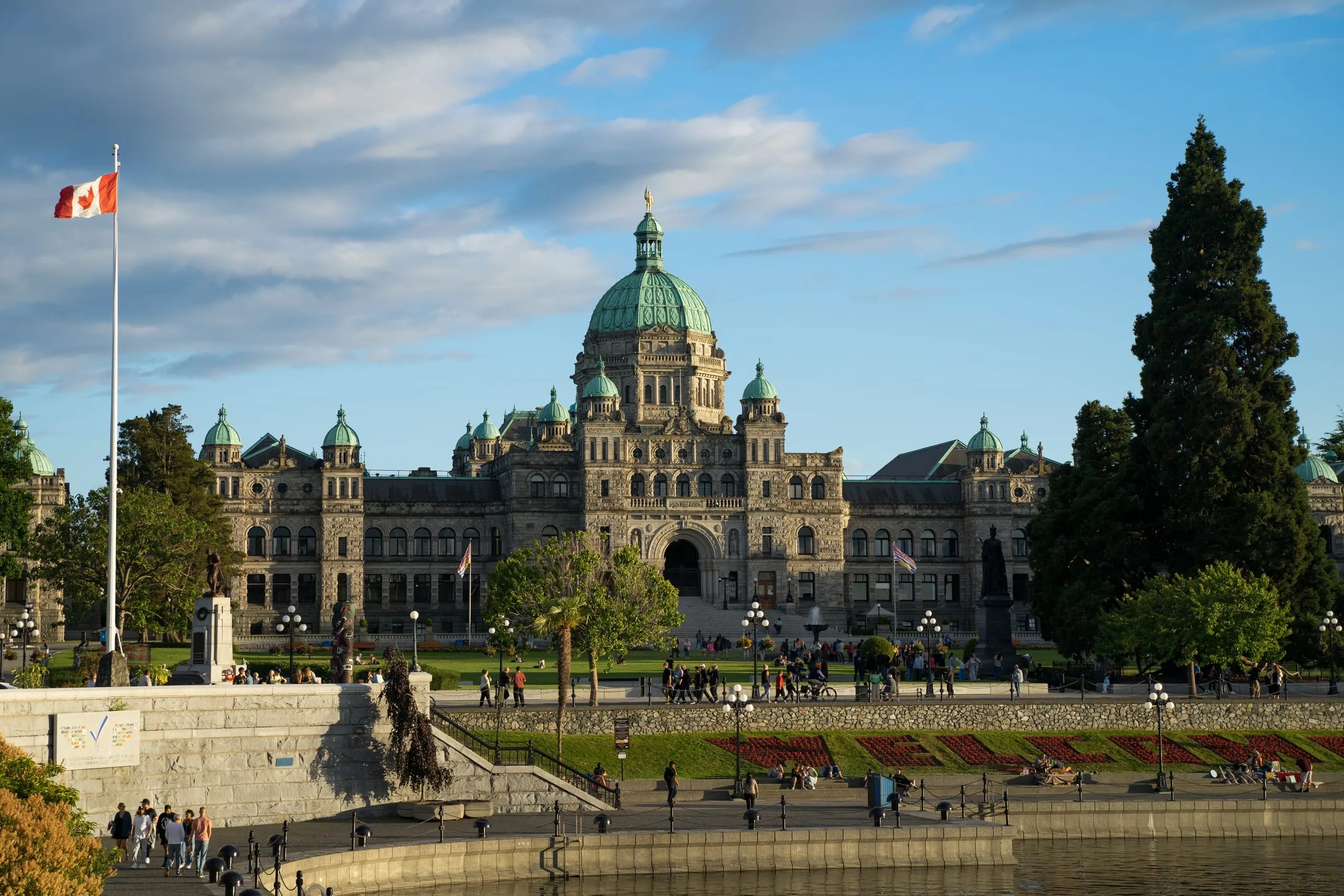 The domed British Columbia Parliament Buildings catch warm evening light above harborfront lawns and a Canadian flag in Victoria.