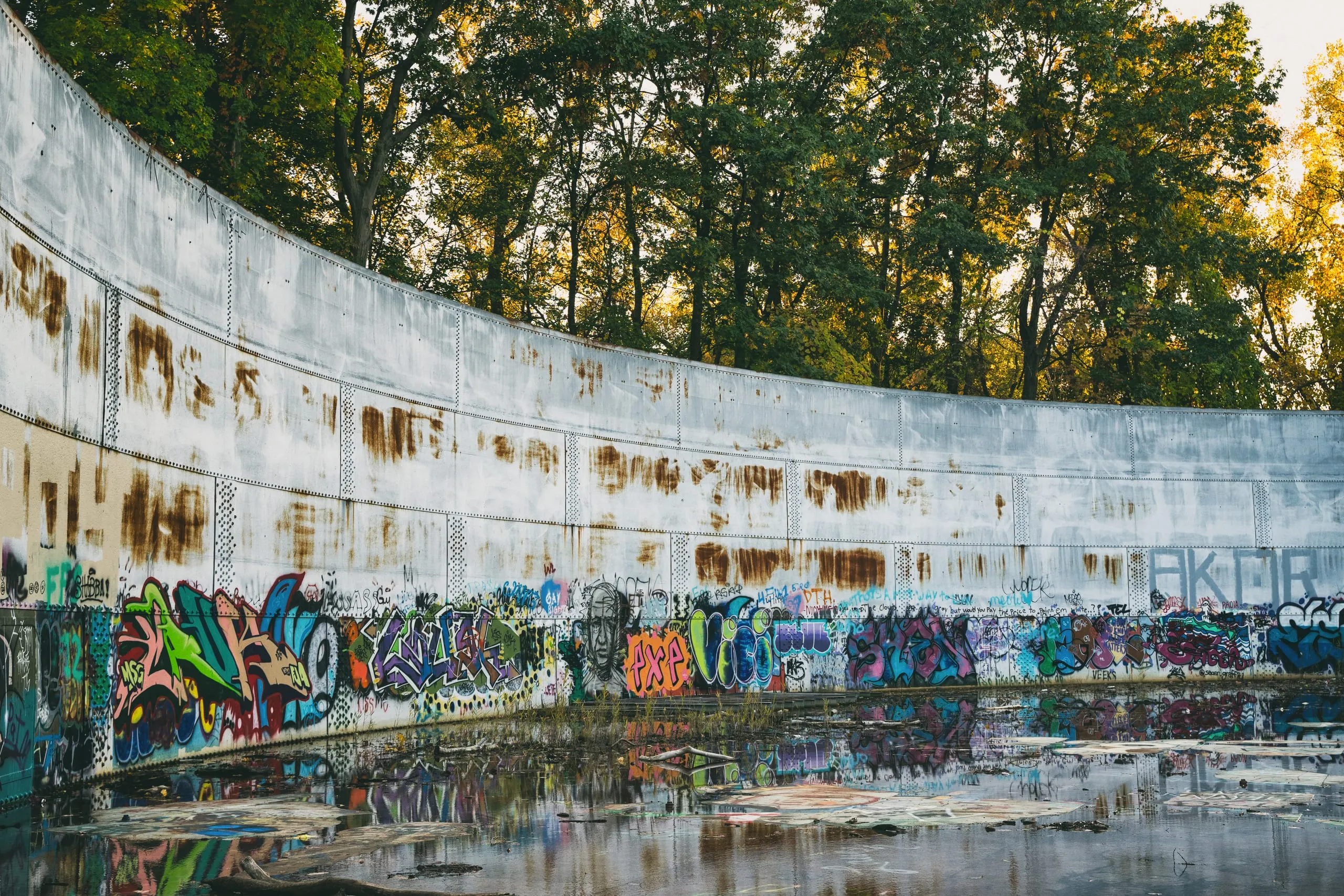 Curved rusted water tank wall covered in graffiti, reflected in a still pool