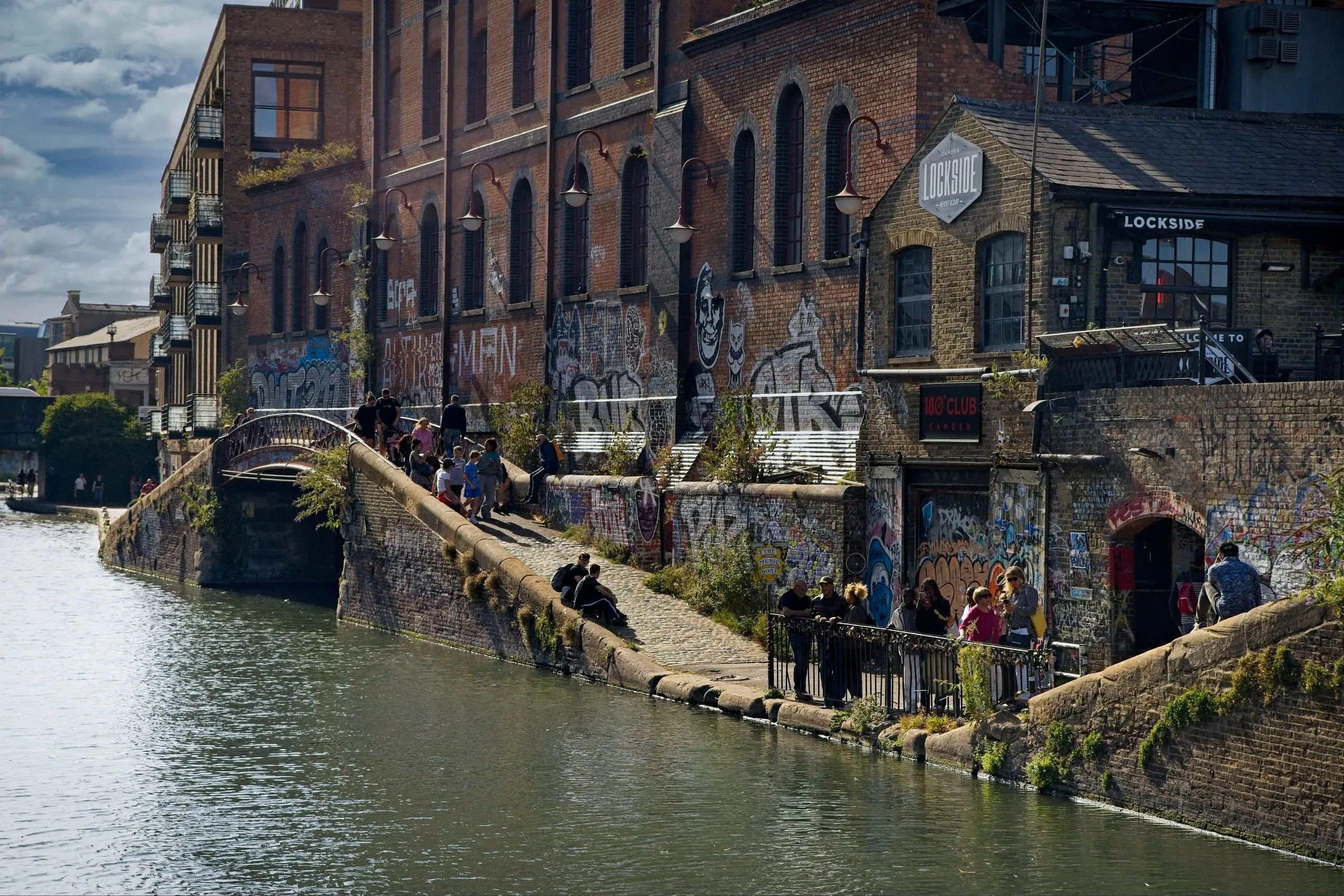 Color photograph of crowds gathered along a graffiti-covered stone towpath and bridge at Camden Lock on Regent's Canal in London, near a building marked 'Lockside'.