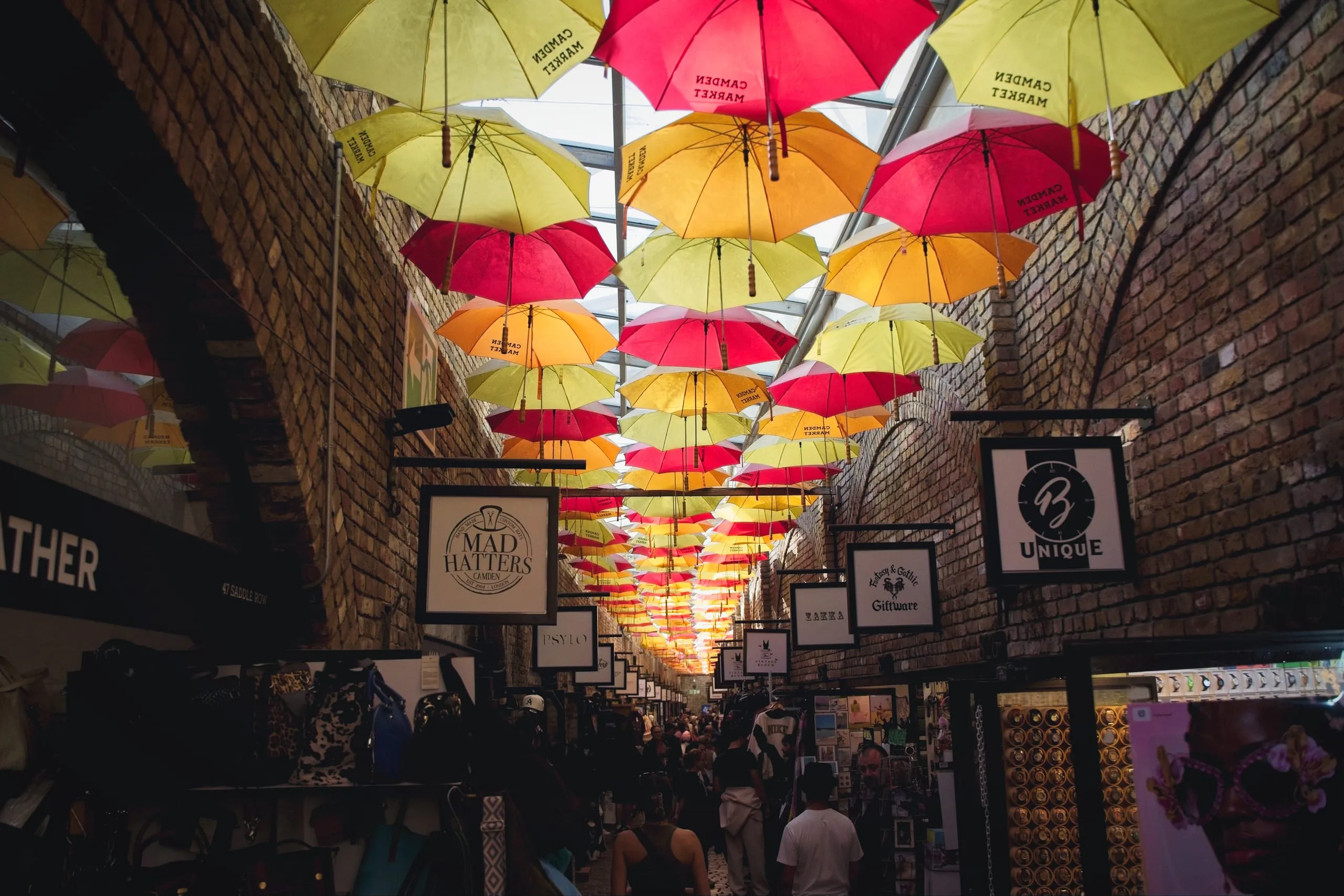 Color photograph of rows of yellow, orange, and red umbrellas printed with 'Camden Market' hanging overhead in a brick-vaulted market arcade in London.
