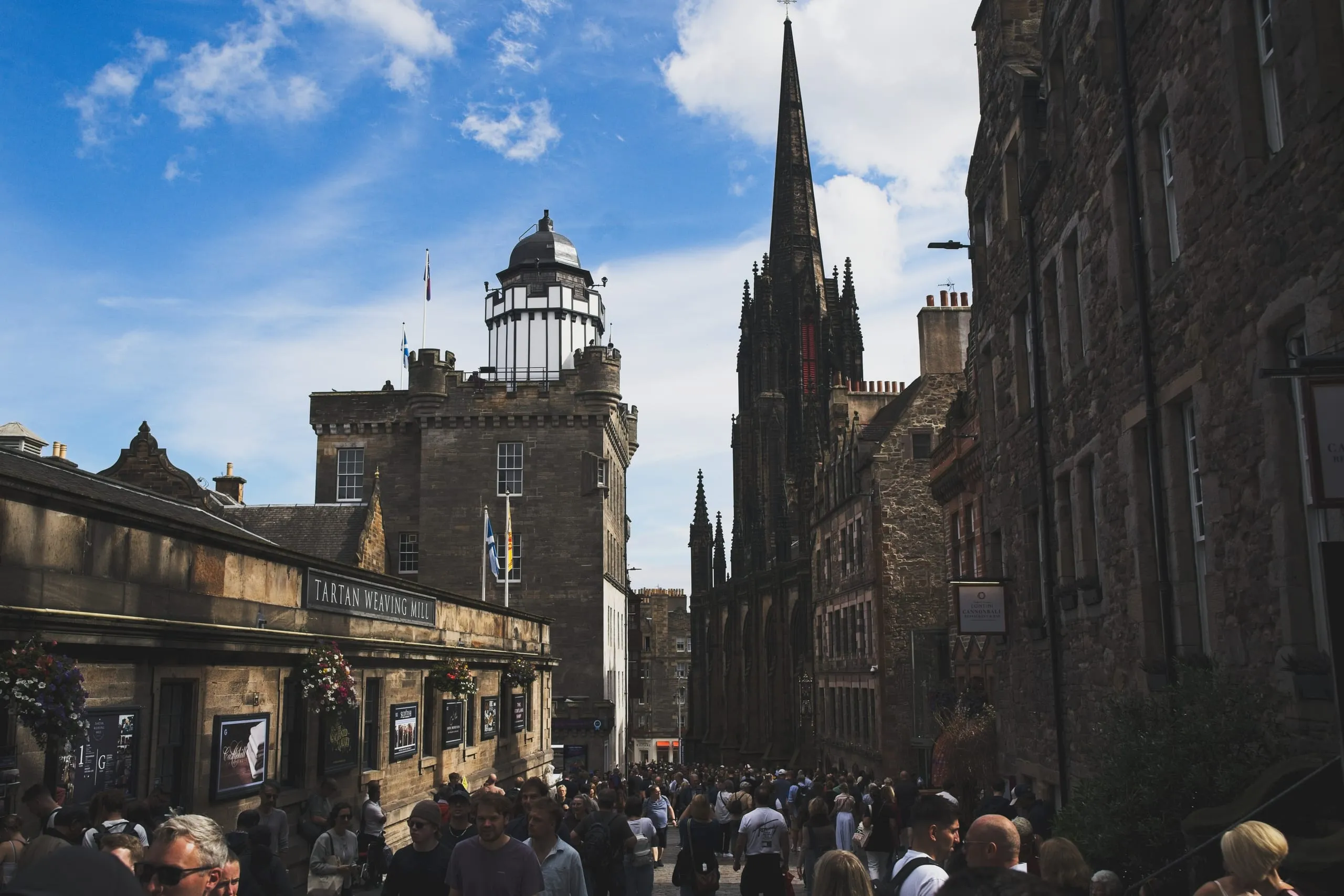 A color photograph of Castlehill on Edinburgh's Royal Mile in daylight, crowds passing the Camera Obscura tower beneath the distant spire of the Hub, with a Tartan Weaving Mill sign along the stone wall.