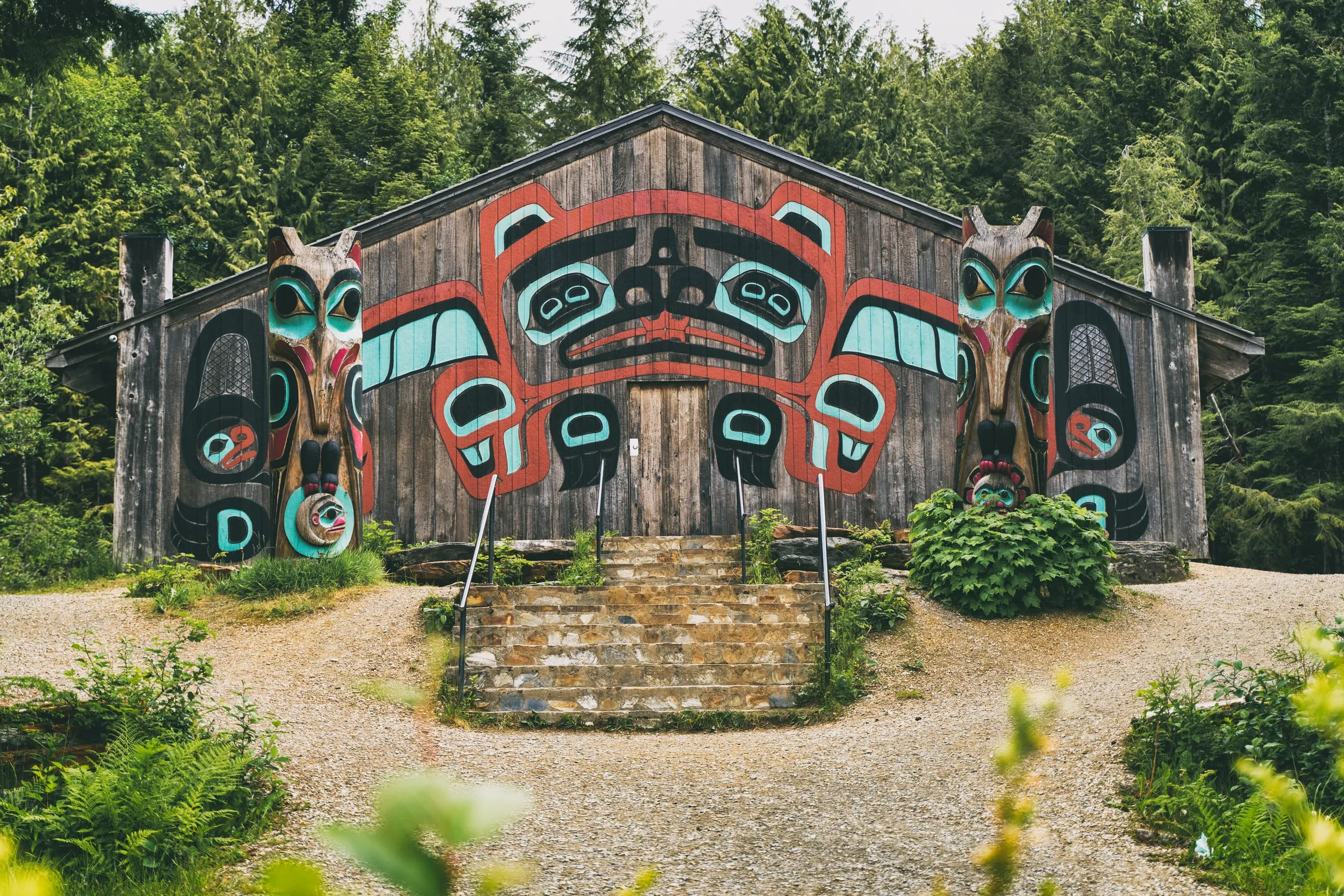 A weathered wood clan house painted with a red and turquoise formline crest, flanked by two carved totem poles, with stone steps up to the door.