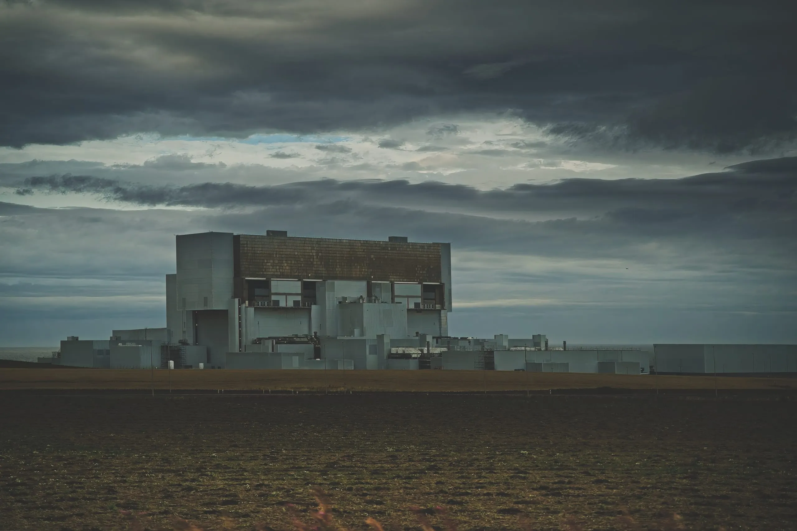 A color photograph of a large industrial power station on the Scottish coast under a dramatic, dark cloud-covered sky, with a plowed field in the foreground.