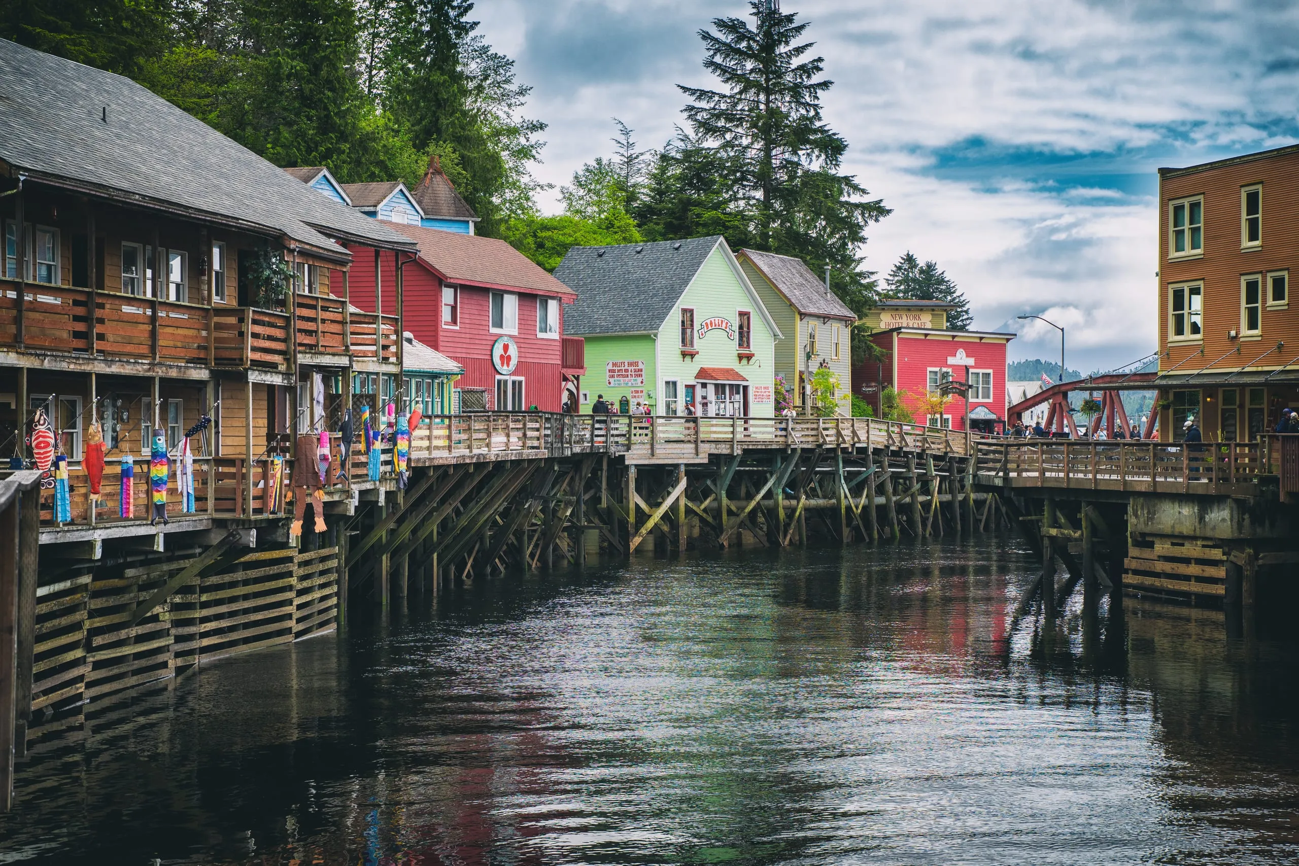 A row of colorful wooden shops on stilts lines a boardwalk above a tidal creek, with decorative fish banners hanging from the railings.