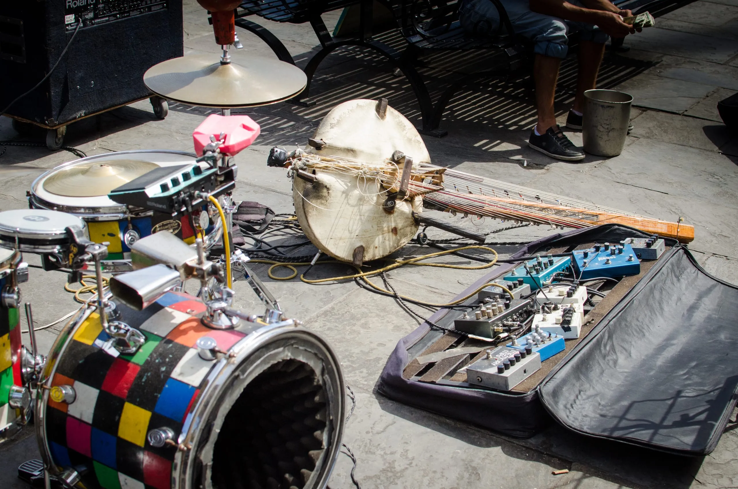 A street musician's gear laid on New Orleans pavement: a multicolored drum kit, cymbal, homemade stringed instrument, and a pedalboard.