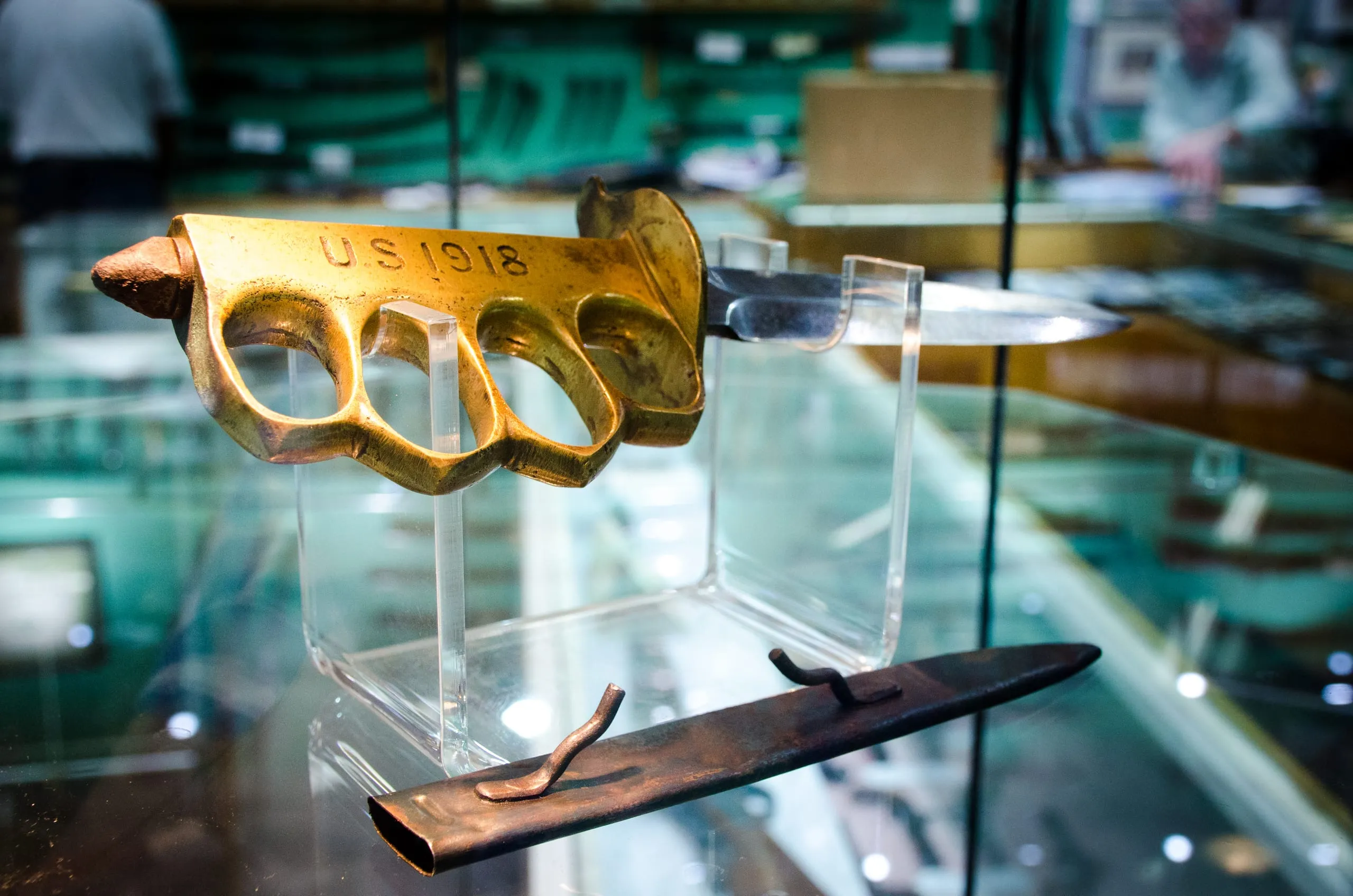 A brass knuckle-handled trench knife stamped "US 1918" on an acrylic stand inside a New Orleans glass display case.