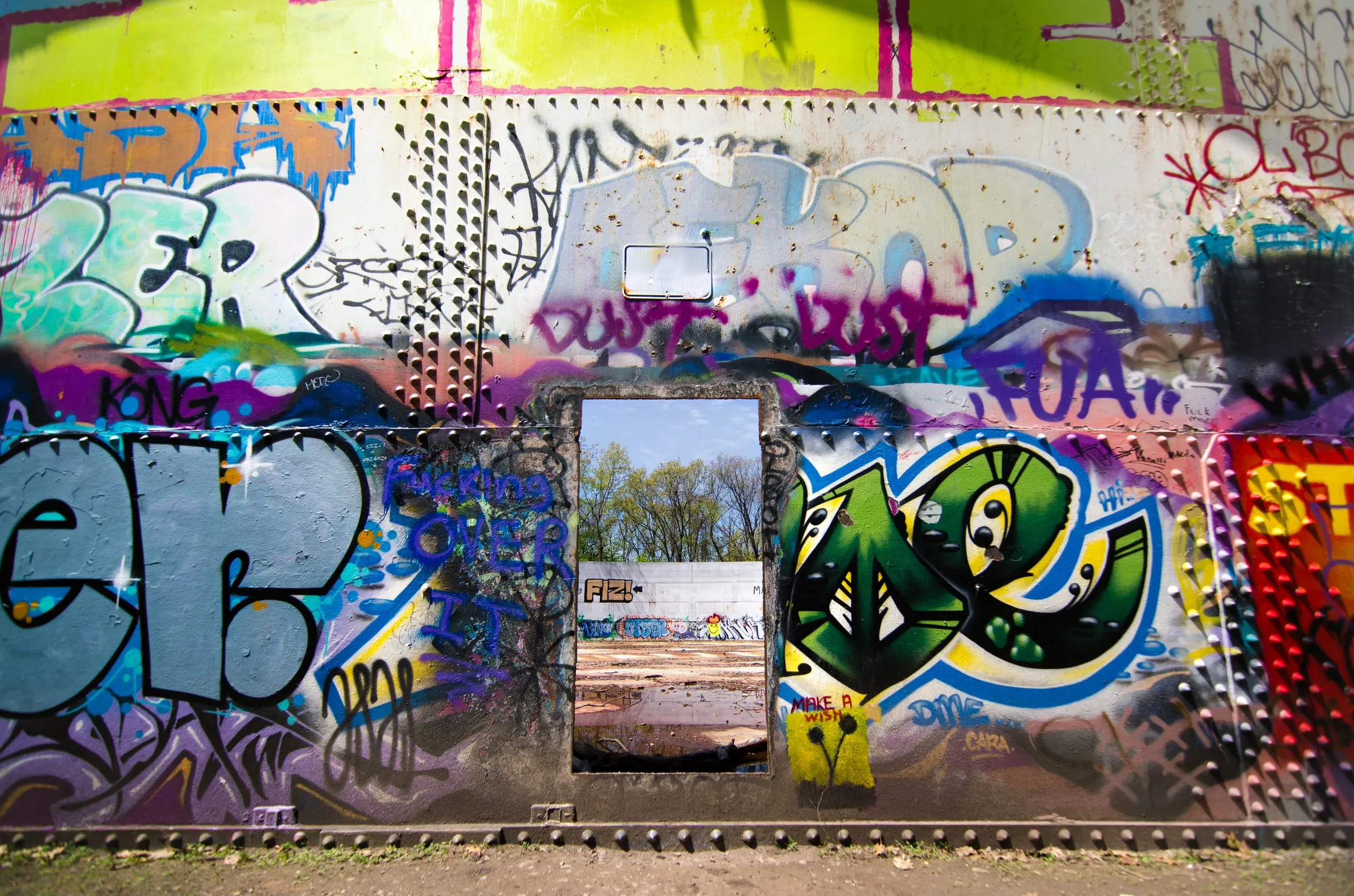 A rectangular doorway cut through a densely graffiti-covered metal wall frames trees and sky in Rochester.