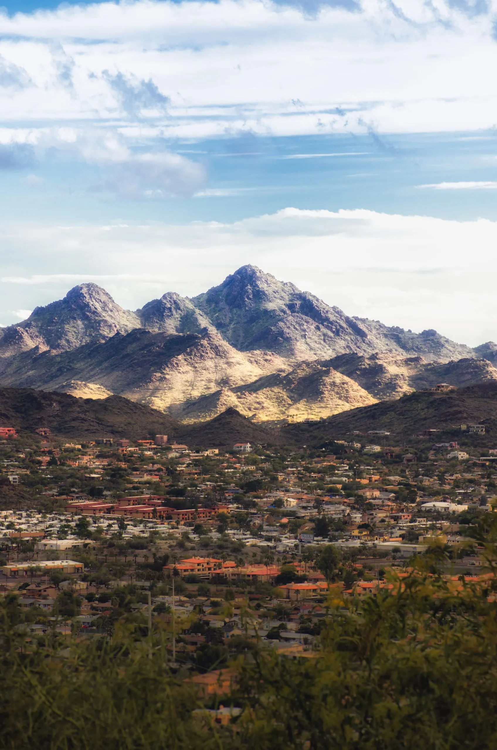 A rugged Arizona mountain range rises above a spread of low desert homes with terracotta roofs and palm trees.