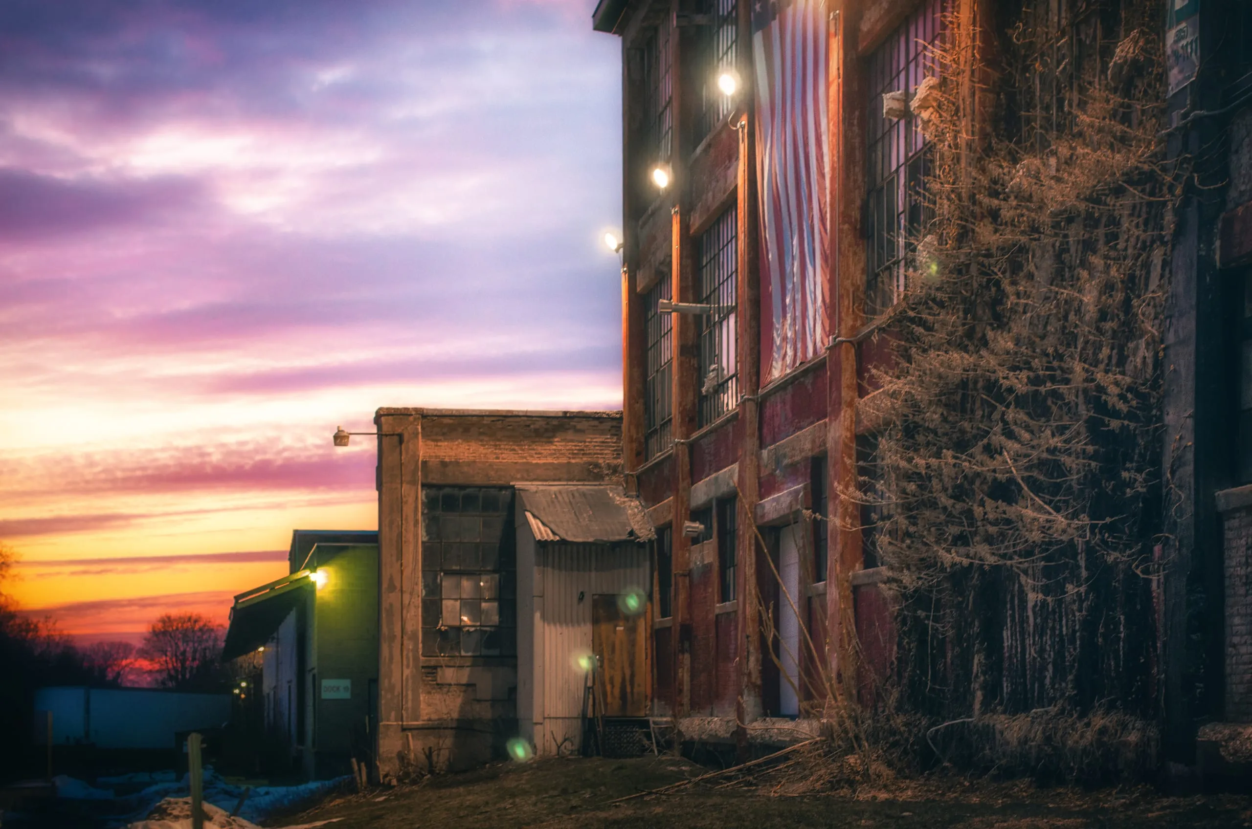 A weathered brick factory with a large American flag hung in its windows glows under a purple-and-orange sunset in Rochester, NY.