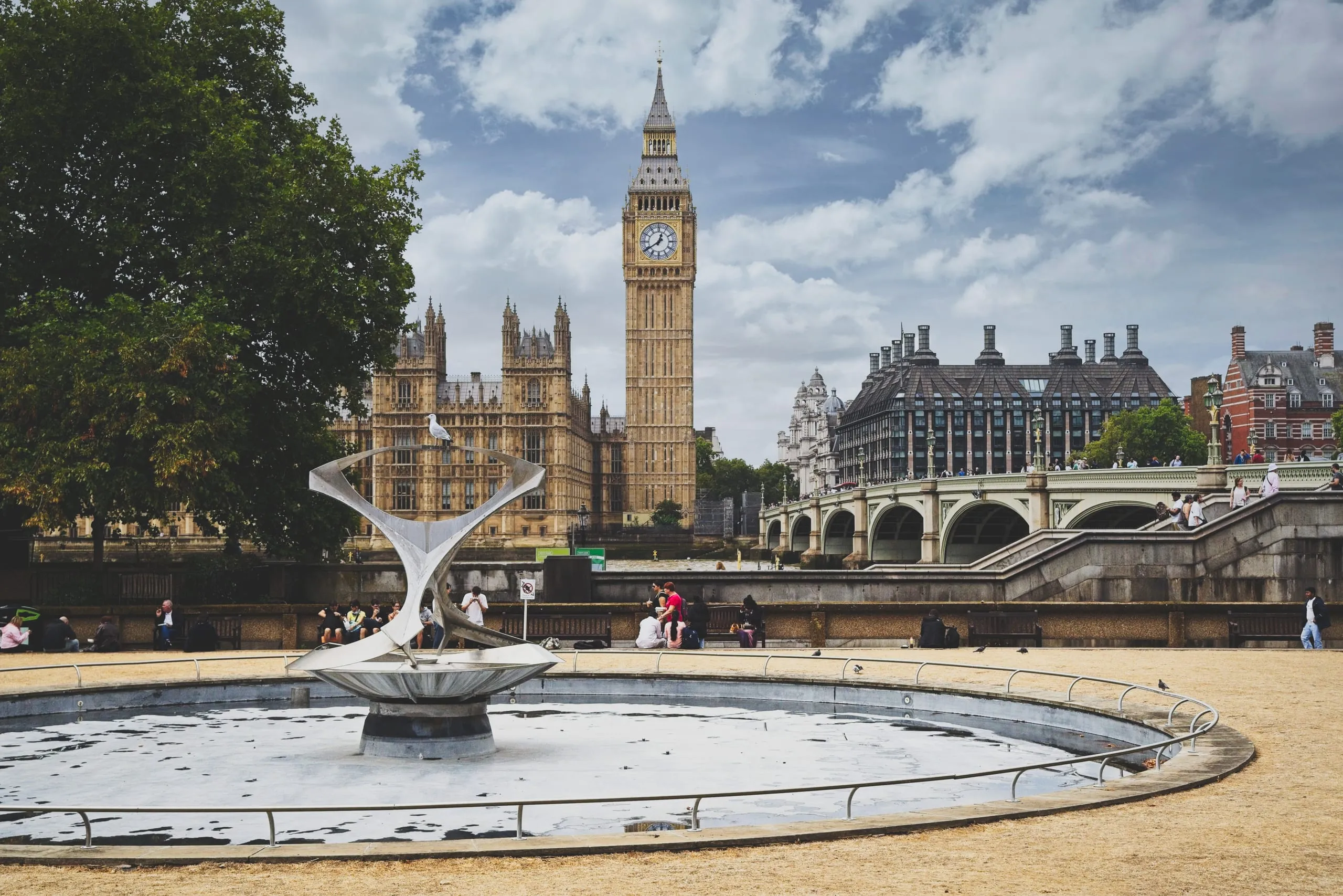 A wide view of an empty circular fountain with a modern steel sculpture in a plaza, with Big Ben and Westminster Bridge visible in the distance, London, England.