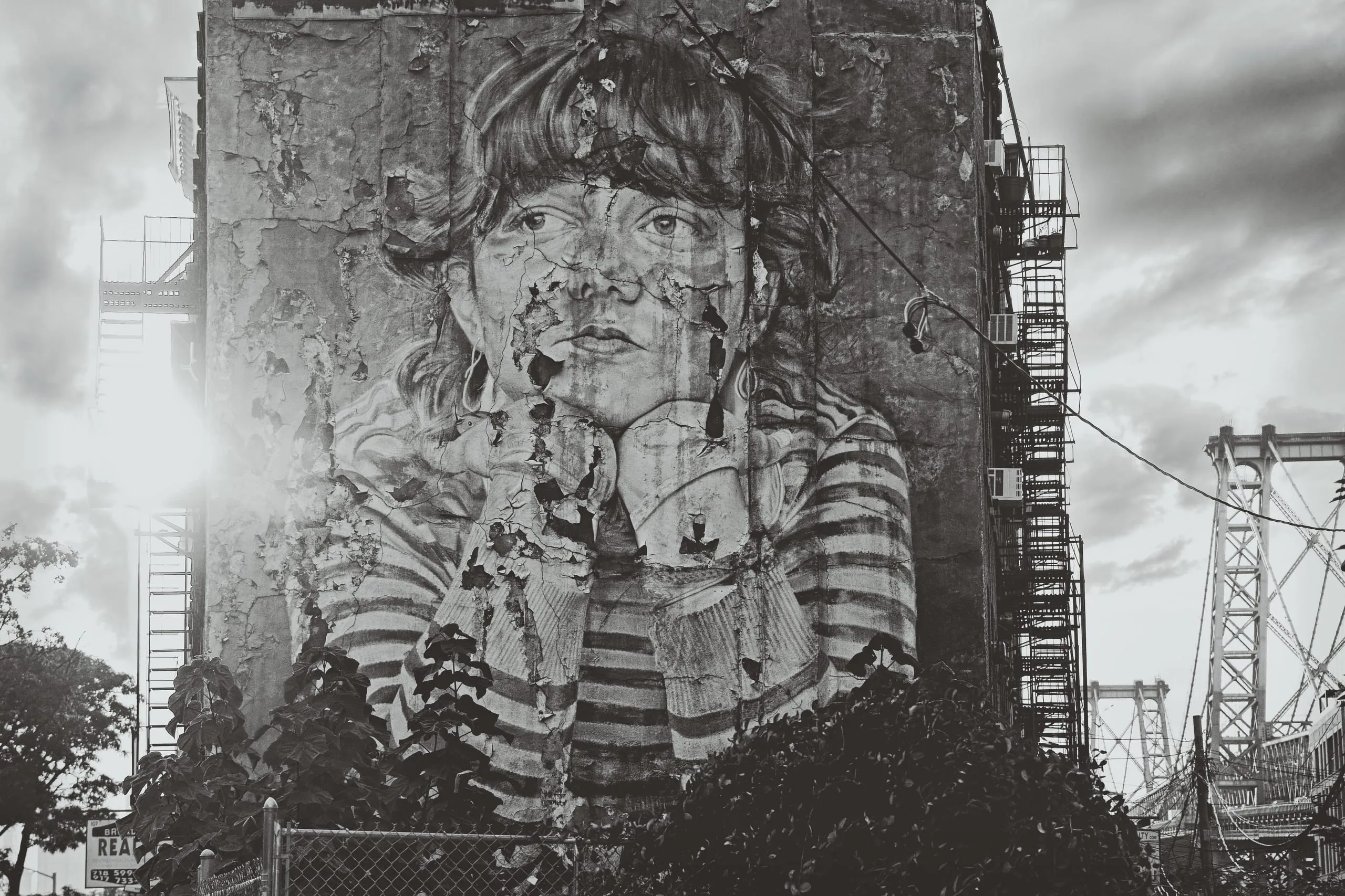 Black and white photograph of a large weathered mural of a woman's face peeling off the brick side of a Williamsburg, Brooklyn building, with fire escapes and the Williamsburg Bridge in the background.