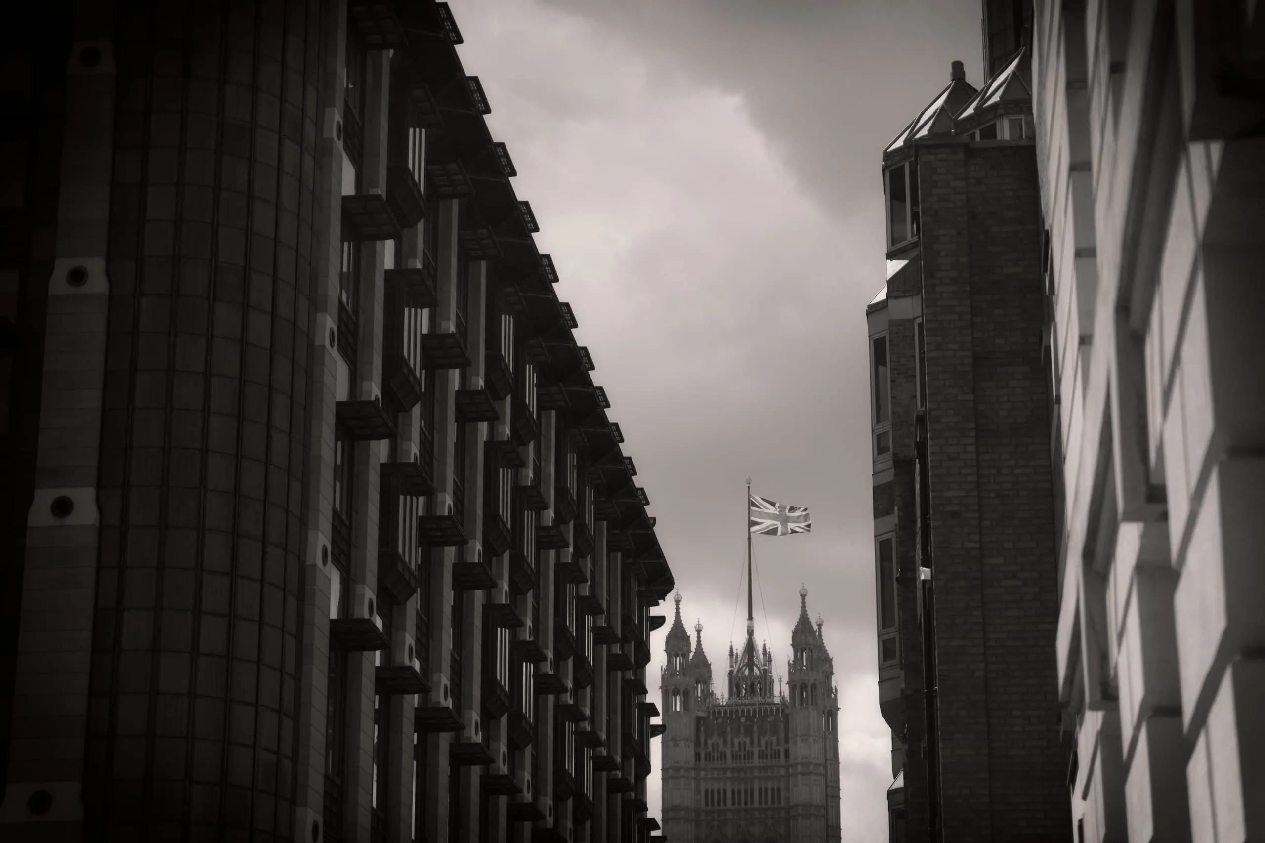 Black and white photograph of the Victoria Tower of the Houses of Parliament, flying a Union Jack flag, framed between two modern buildings beneath an overcast sky, London, England.