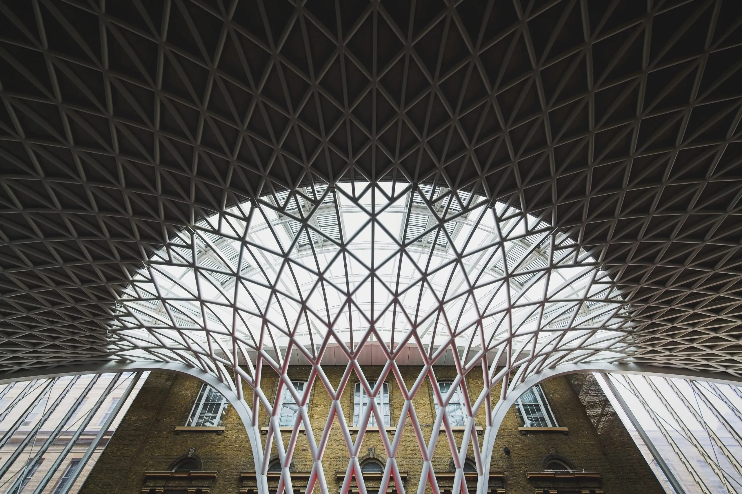 A photograph looking straight up at the lattice funnel roof of King's Cross station's Western Concourse in London, with a brick building visible through the glass at its base.
