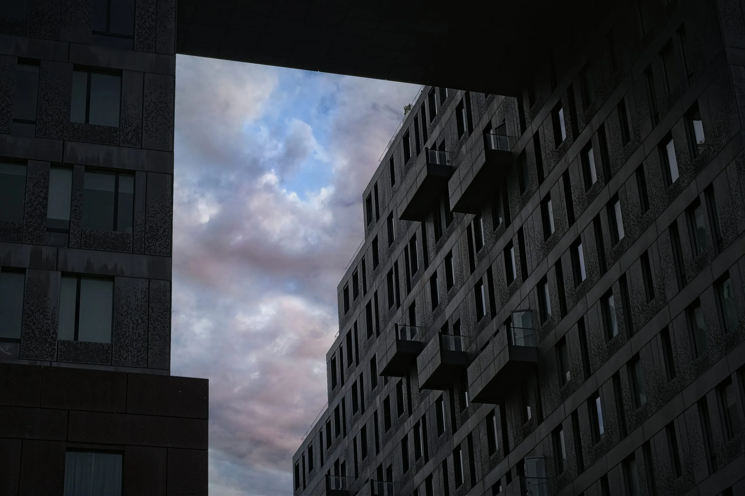 A color photograph of a narrow sliver of pink and blue dusk sky framed between two tall dark apartment buildings in Brooklyn.