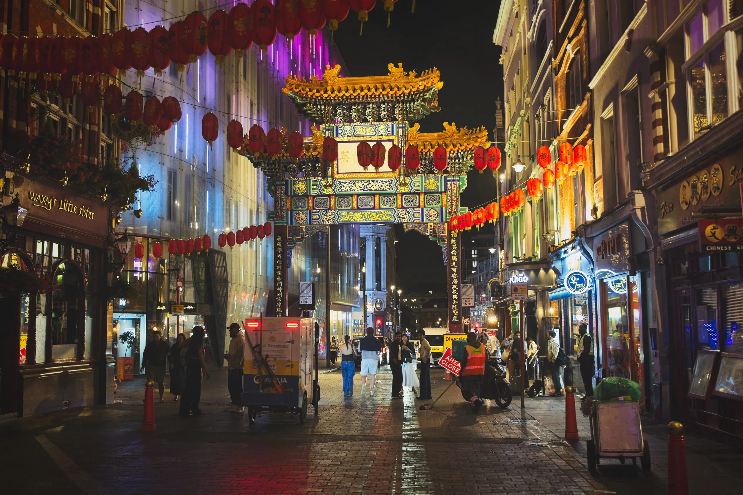 Photograph of the ornate gold-roofed Chinatown gate arching over a lantern-strung street at night, with street cleaners and pedestrians beneath it, London, England.