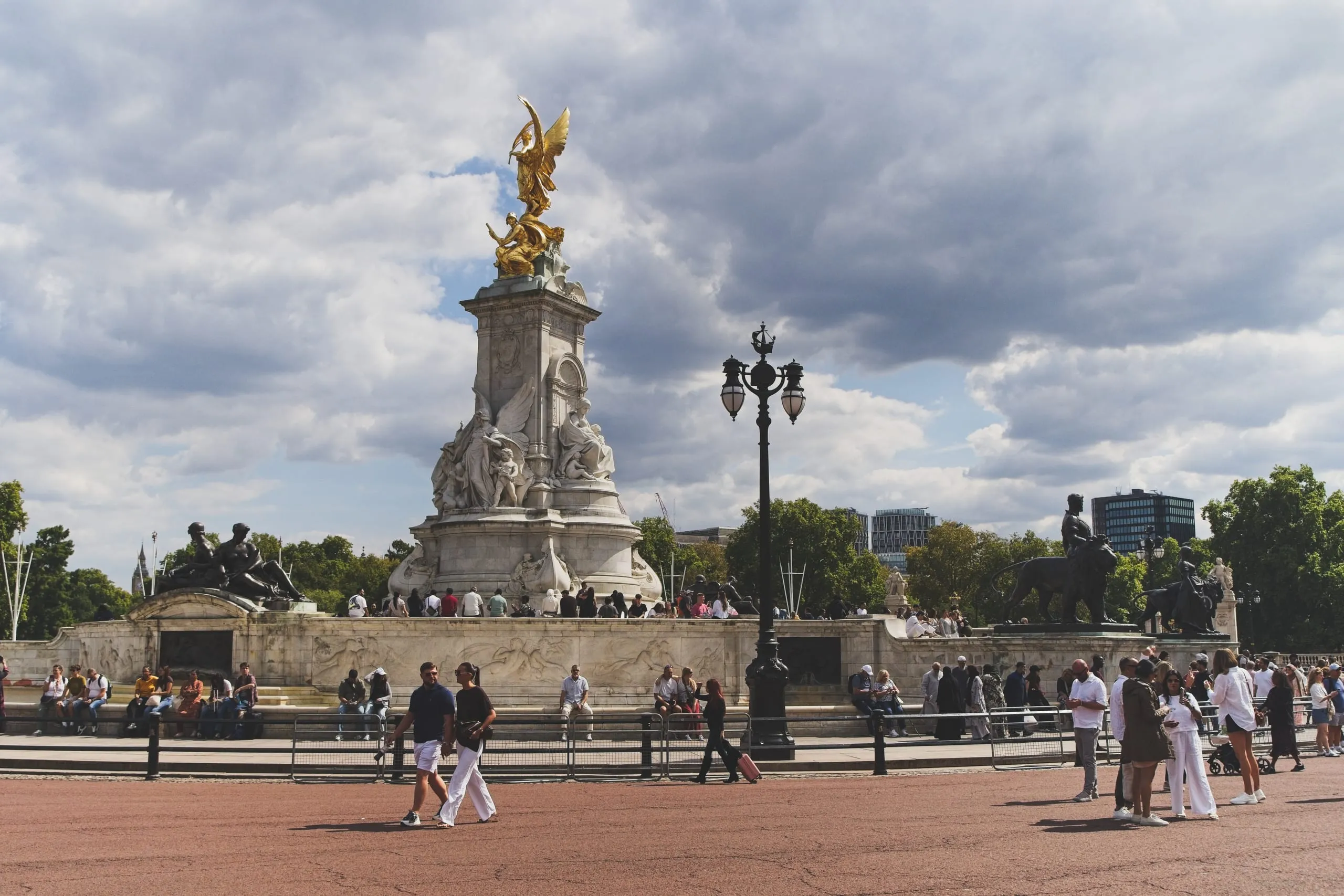 A photograph of the gilded winged Victory statue atop the Victoria Memorial outside Buckingham Palace in London, with tourists gathered around the marble base under a cloudy sky.