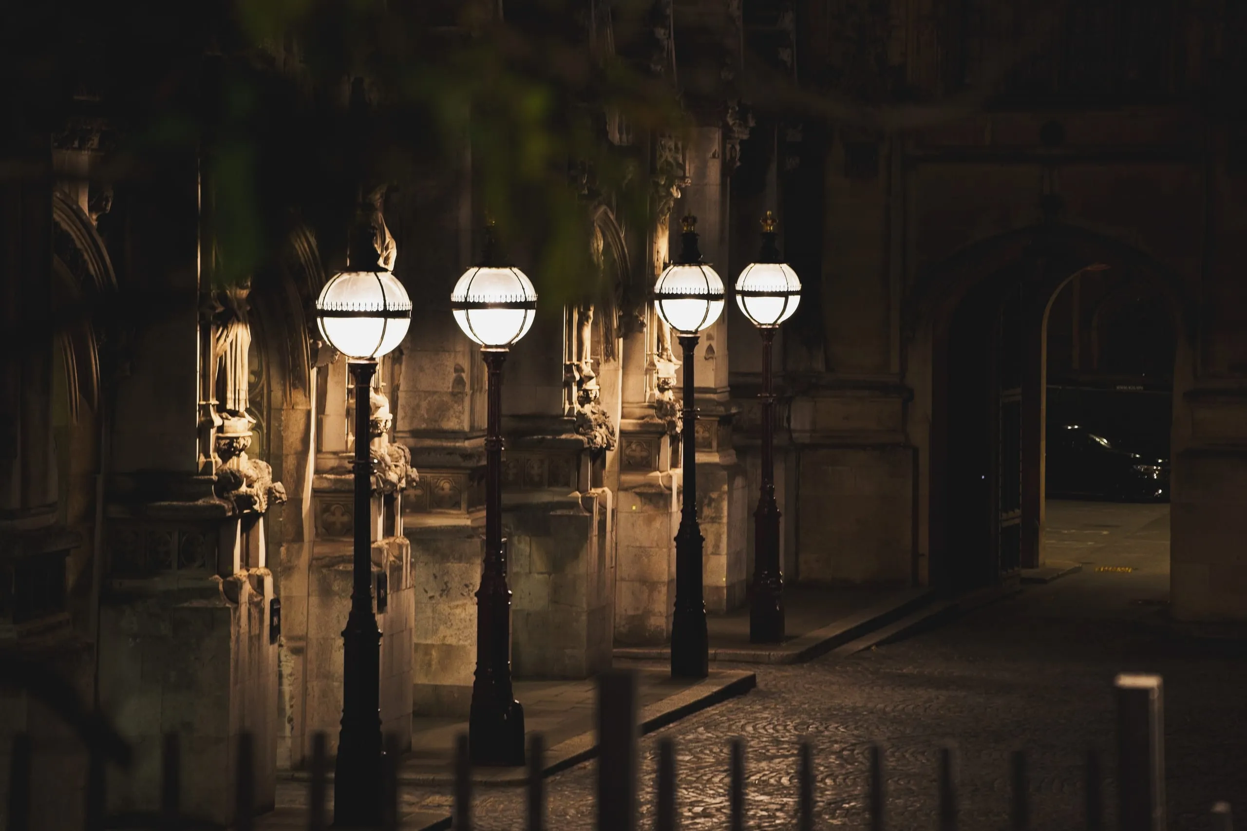A night photograph of a row of glowing globe lampposts along a cobblestone path beside a Gothic stone building, likely near the Houses of Parliament in London, England.