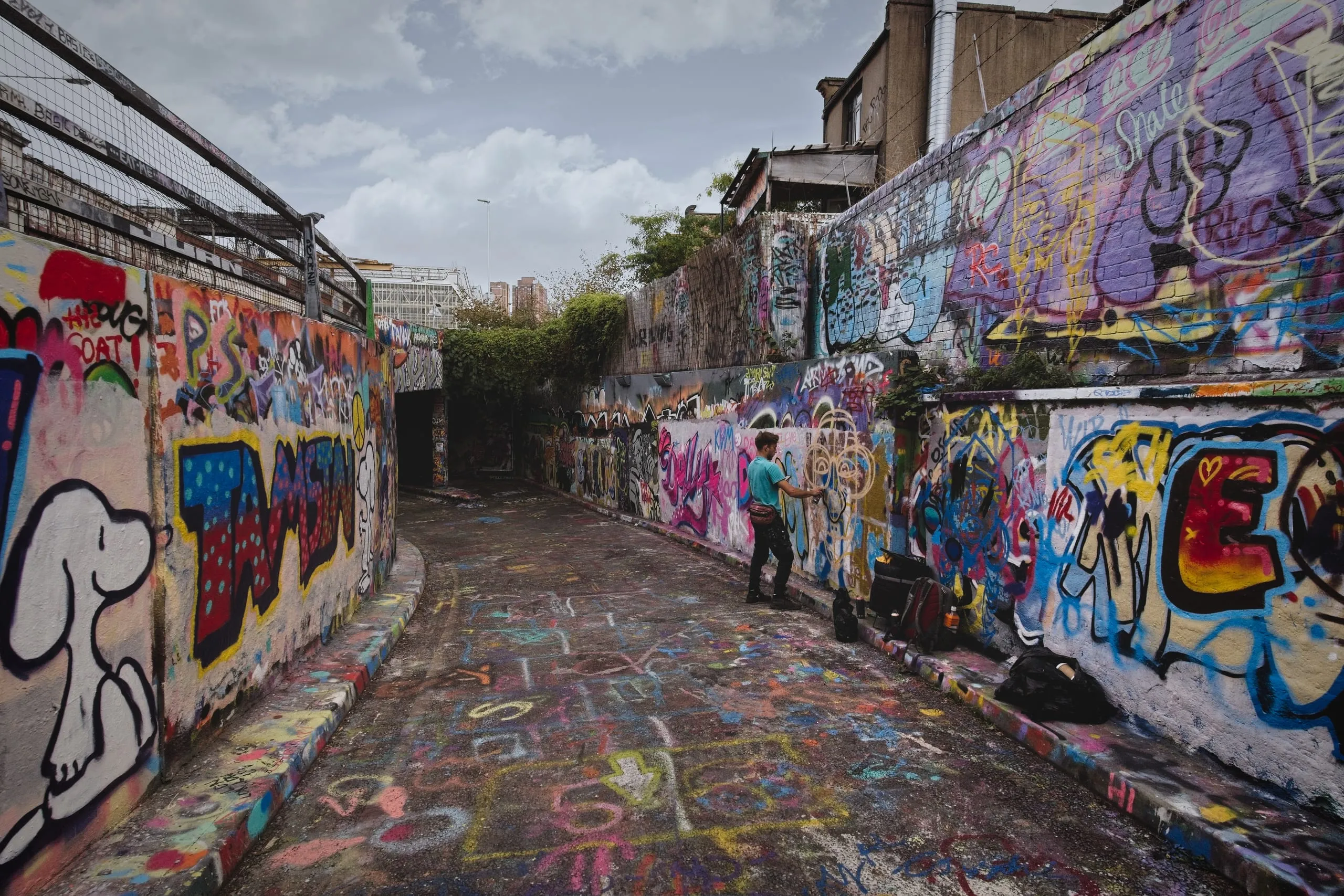 Photograph of an outdoor graffiti-covered alley in London under a cloudy sky, with a man in a teal shirt actively spray-painting a wall, cans and bags set on the curb beside him.