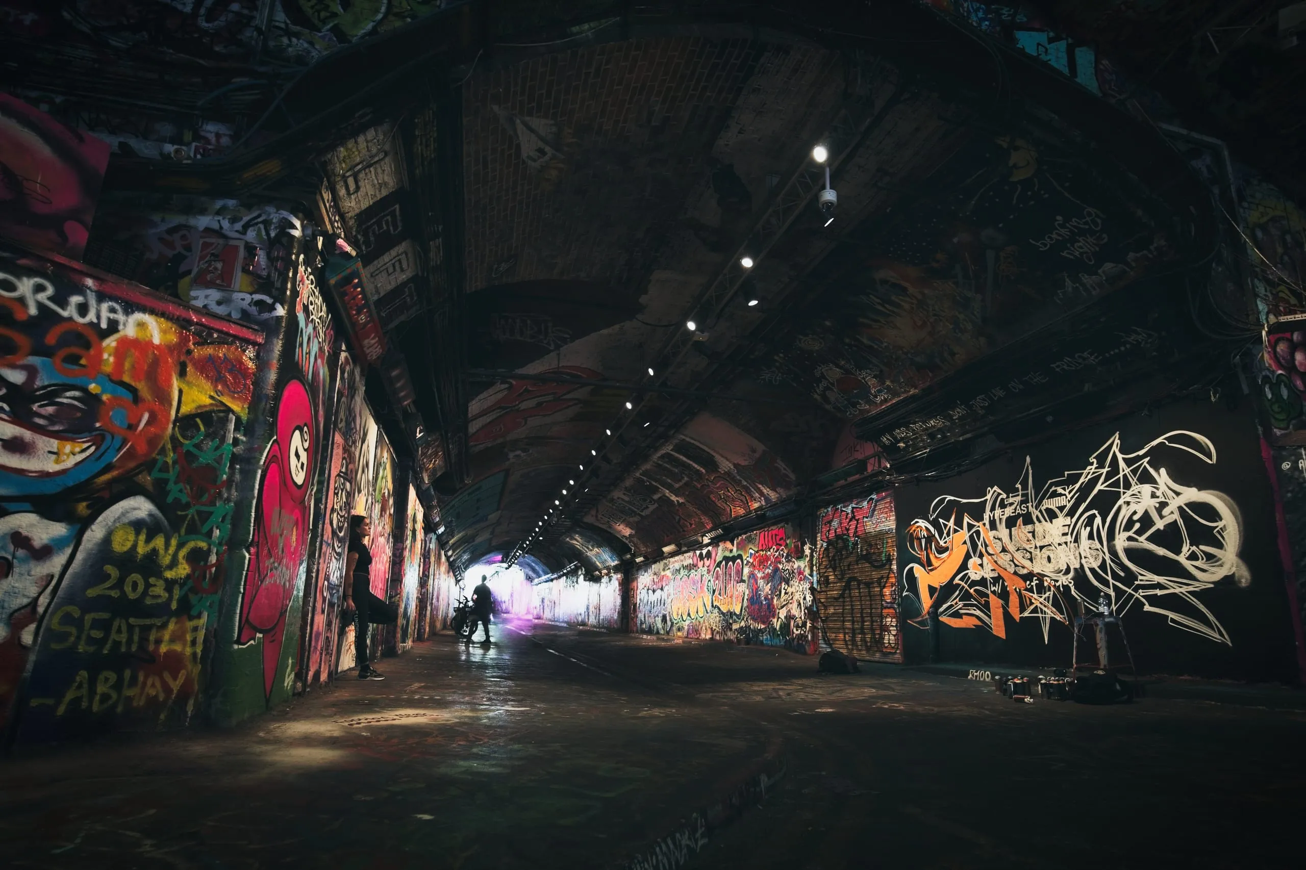 Photograph of a long, vaulted brick tunnel in London with every wall covered in colorful graffiti, two people silhouetted in a shaft of light near the far end.