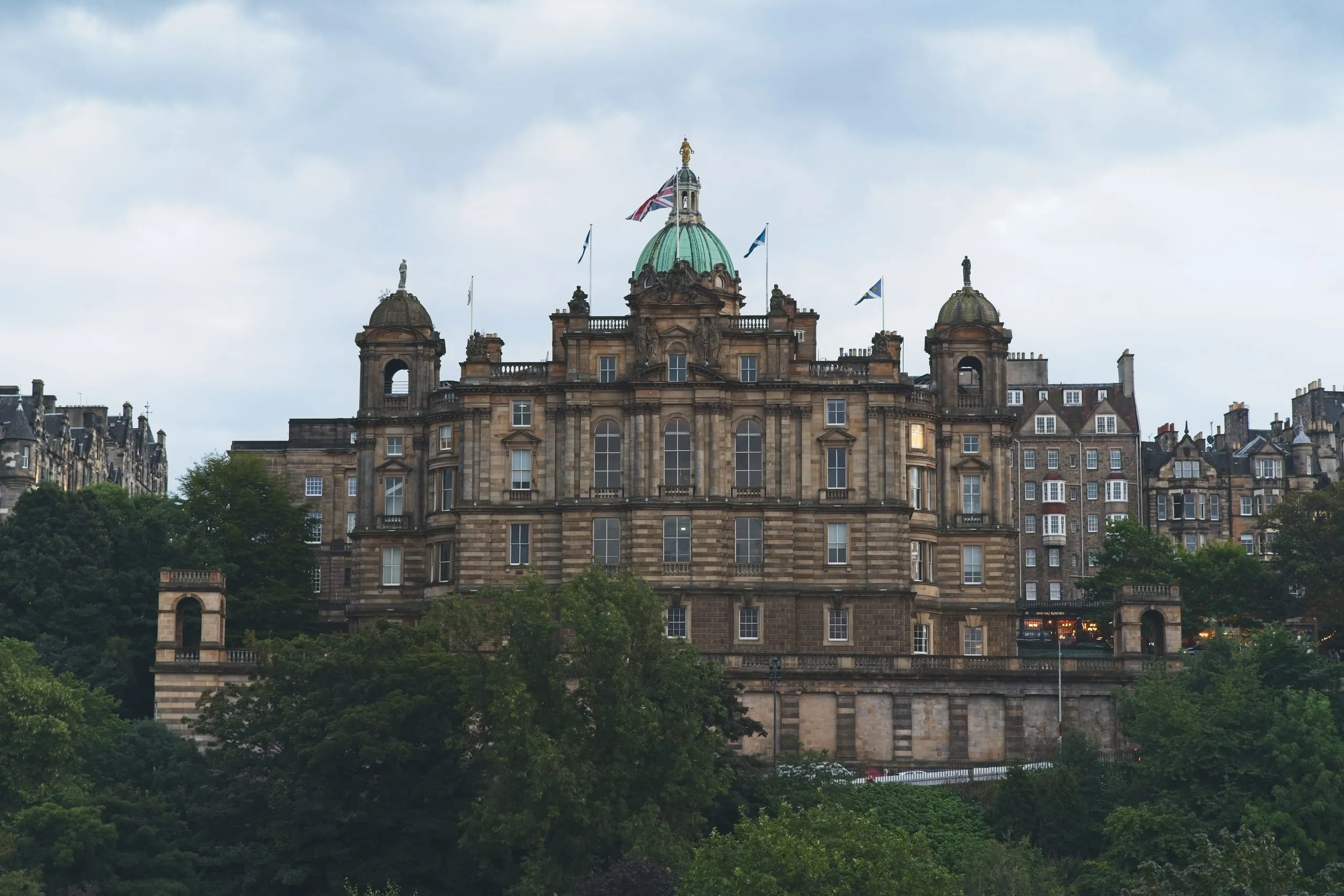 A color photograph of a grand sandstone building with a green copper dome and flags, resembling the Bank of Scotland headquarters on The Mound, overlooking Edinburgh's Old Town.