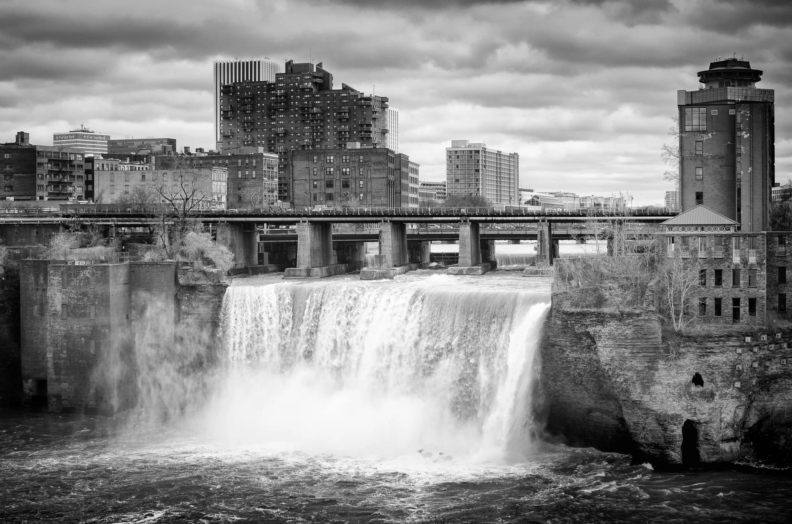 Black and white photograph of High Falls waterfall with the Rochester skyline under a storm sky