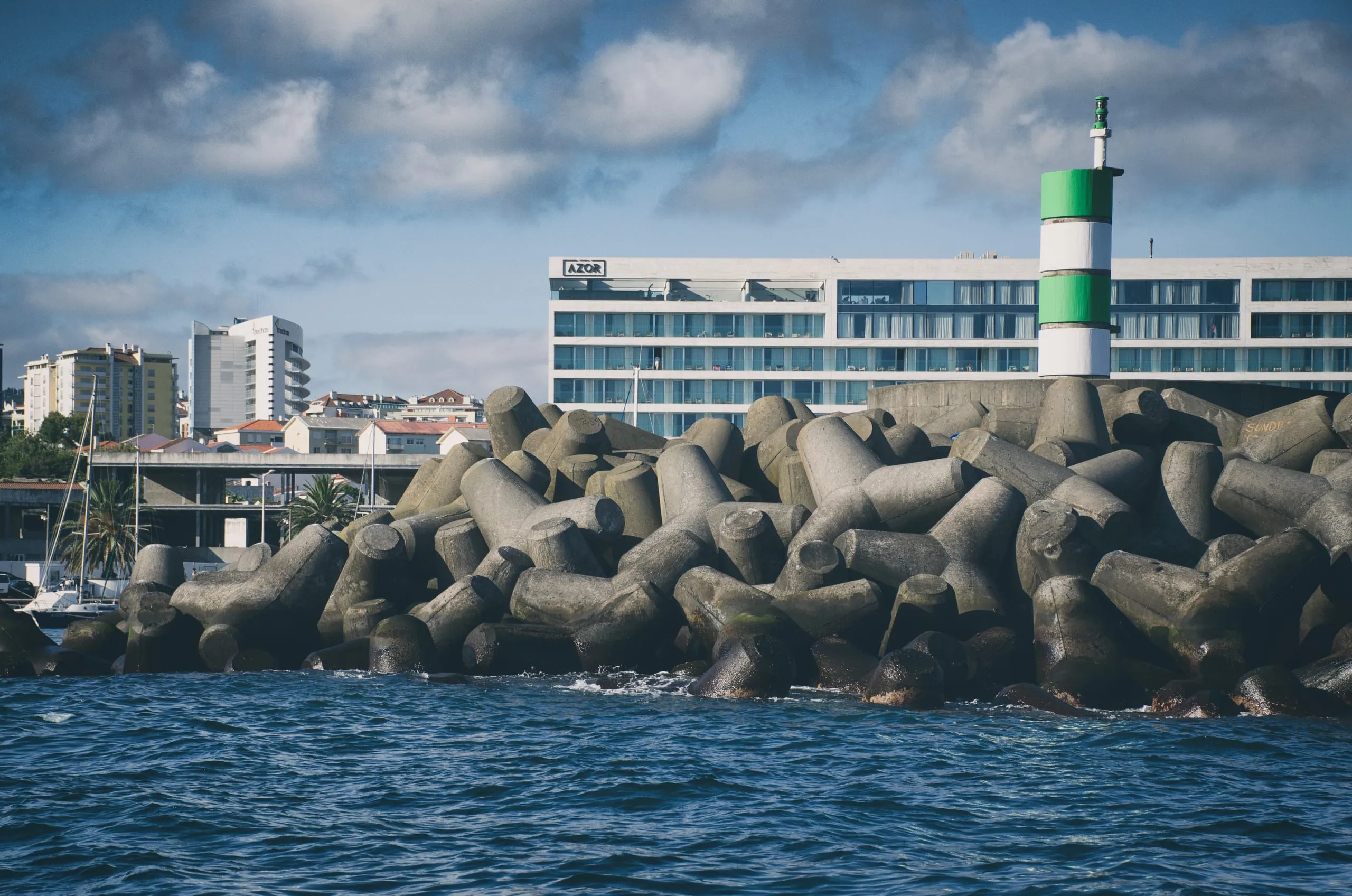 A harbor breakwater piled with concrete tetrapods topped by a green-and-white channel light, with a large waterfront hotel and marina behind.