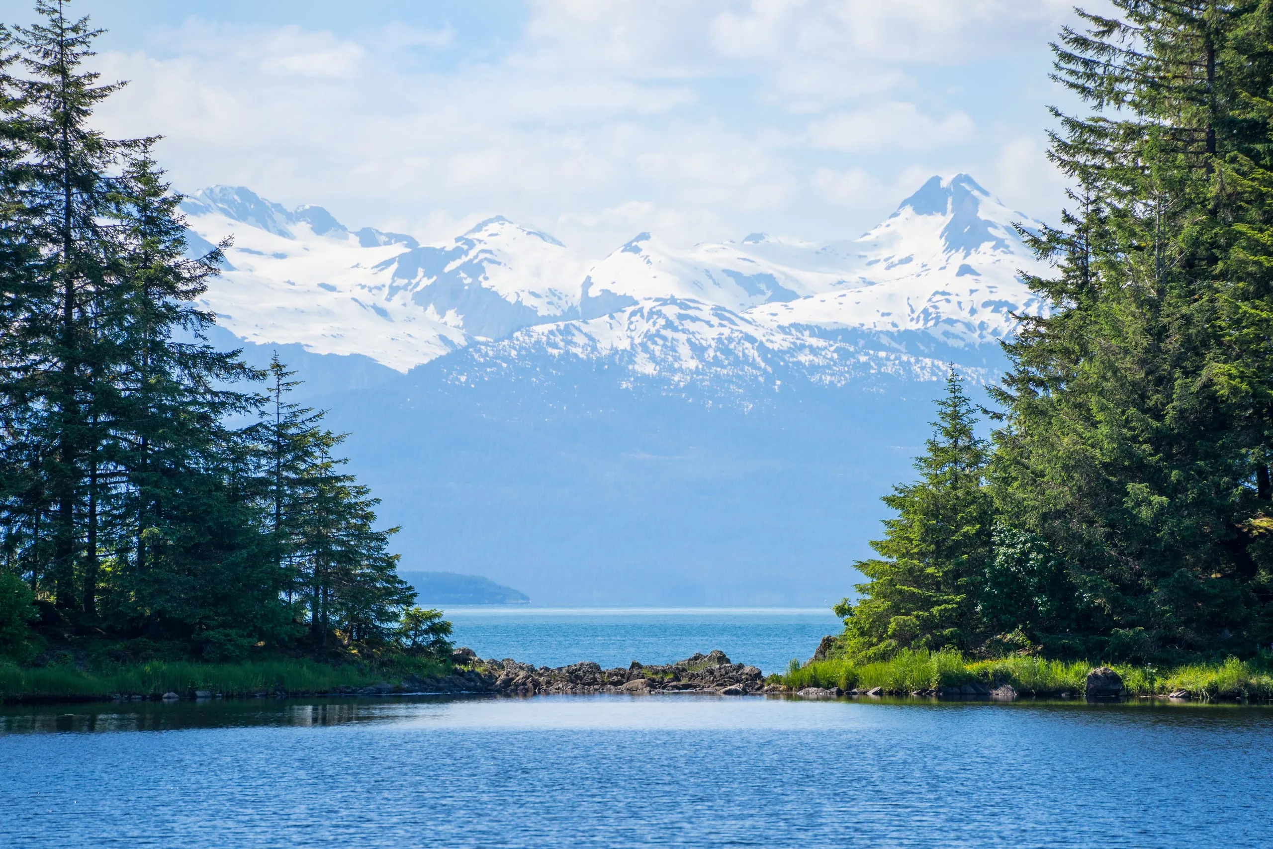 A calm spruce-framed lagoon opens onto a channel with a rocky spit, backed by a hazy snow-capped mountain range.