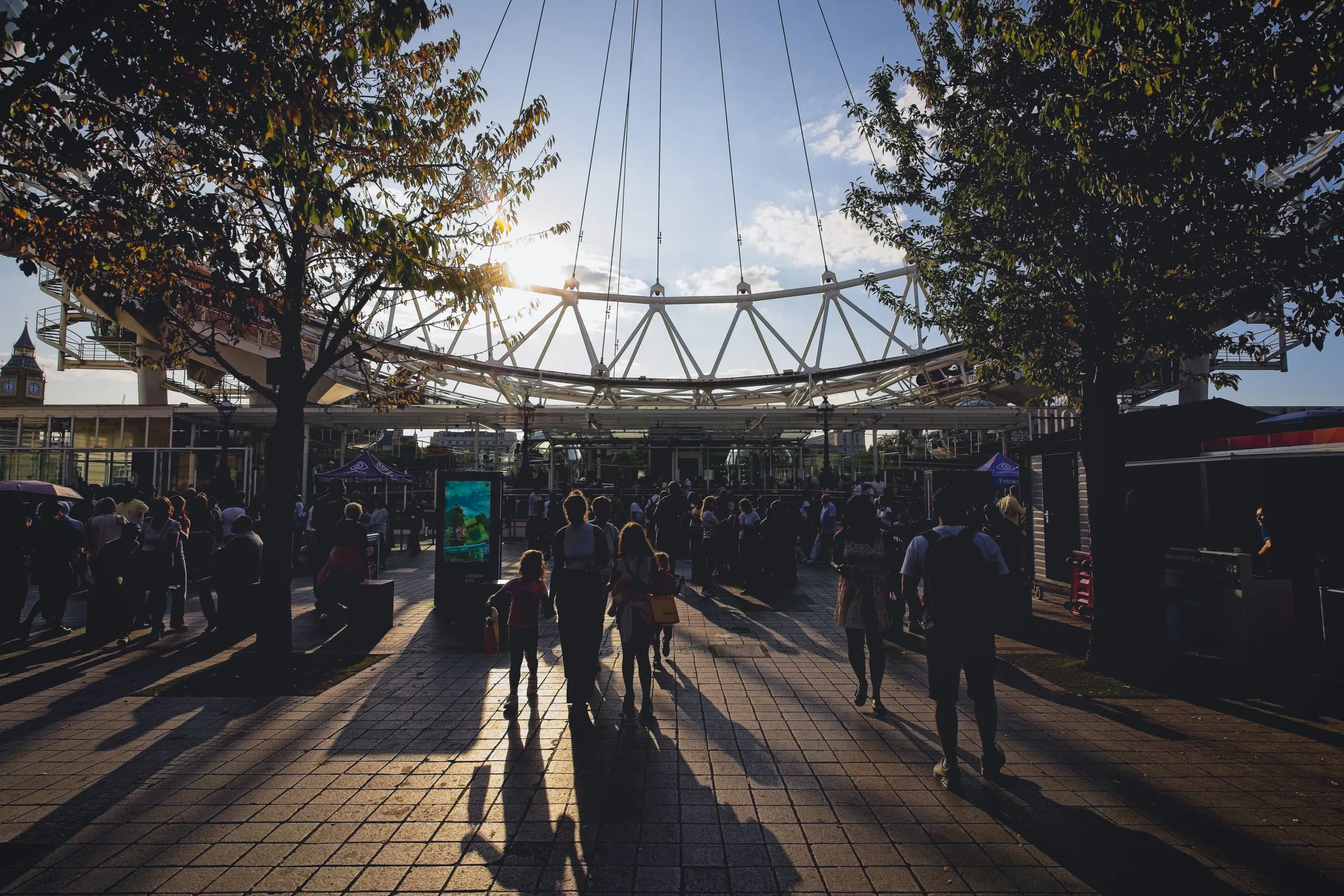 Photograph of tourists in silhouette crossing a sunlit plaza beneath the London Eye, with Big Ben visible in the distance, London, England.