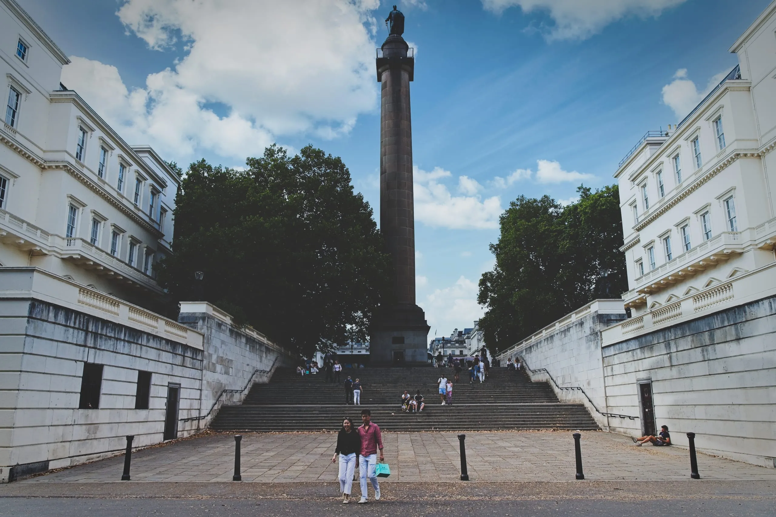 A photograph of the Duke of York Column in London seen from the bottom of its wide staircase, flanked by white Regency buildings with tourists on the steps.