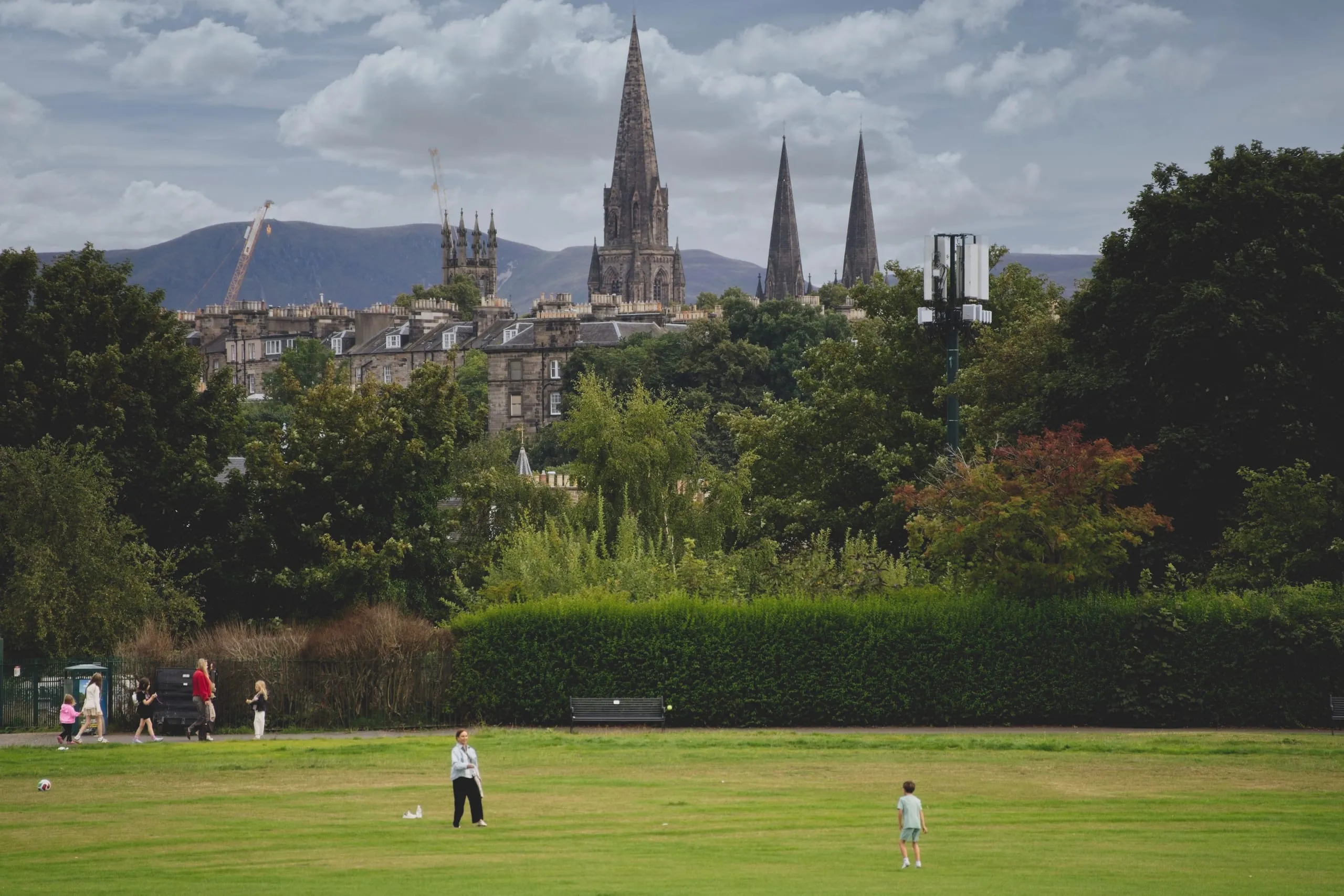 A color photograph of Edinburgh's Meadows park, people playing on the open lawn with the Gothic spires of St Mary's Cathedral and the Pentland Hills rising beyond the rooftops.