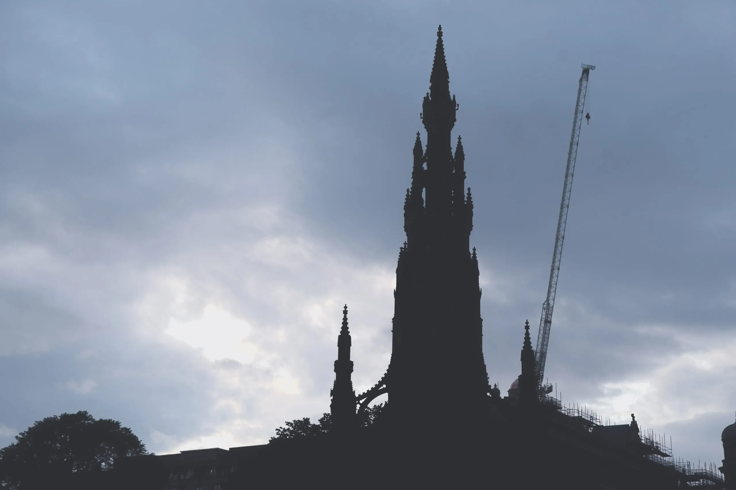 A color photograph of the Scott Monument in Edinburgh silhouetted against a cloudy sky, with a construction crane standing beside it.