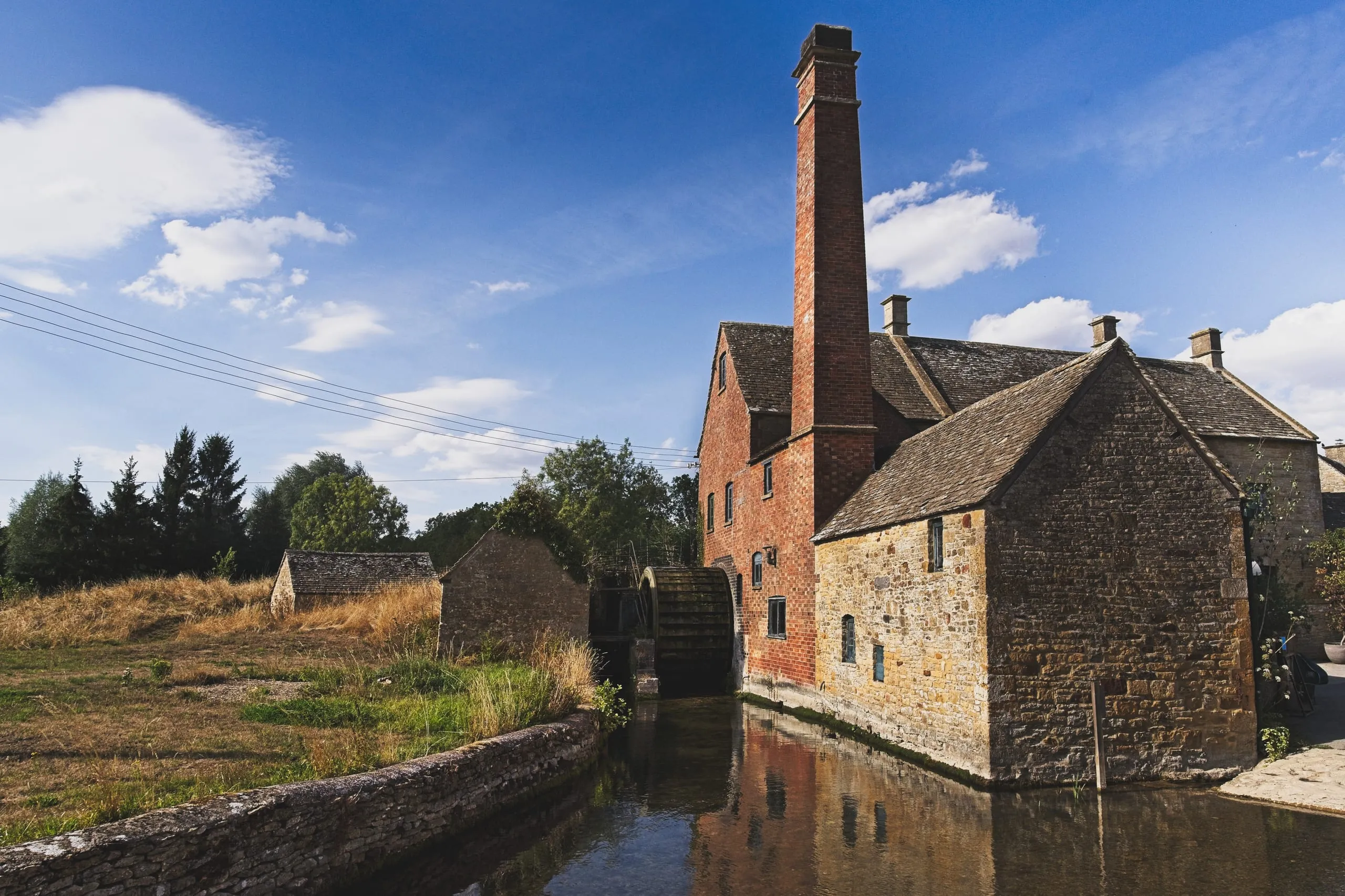 A photograph of the Old Mill in Lower Slaughter, the Cotswolds, England, showing a tall red-brick chimney, a wooden waterwheel, and its reflection in the adjoining stream.