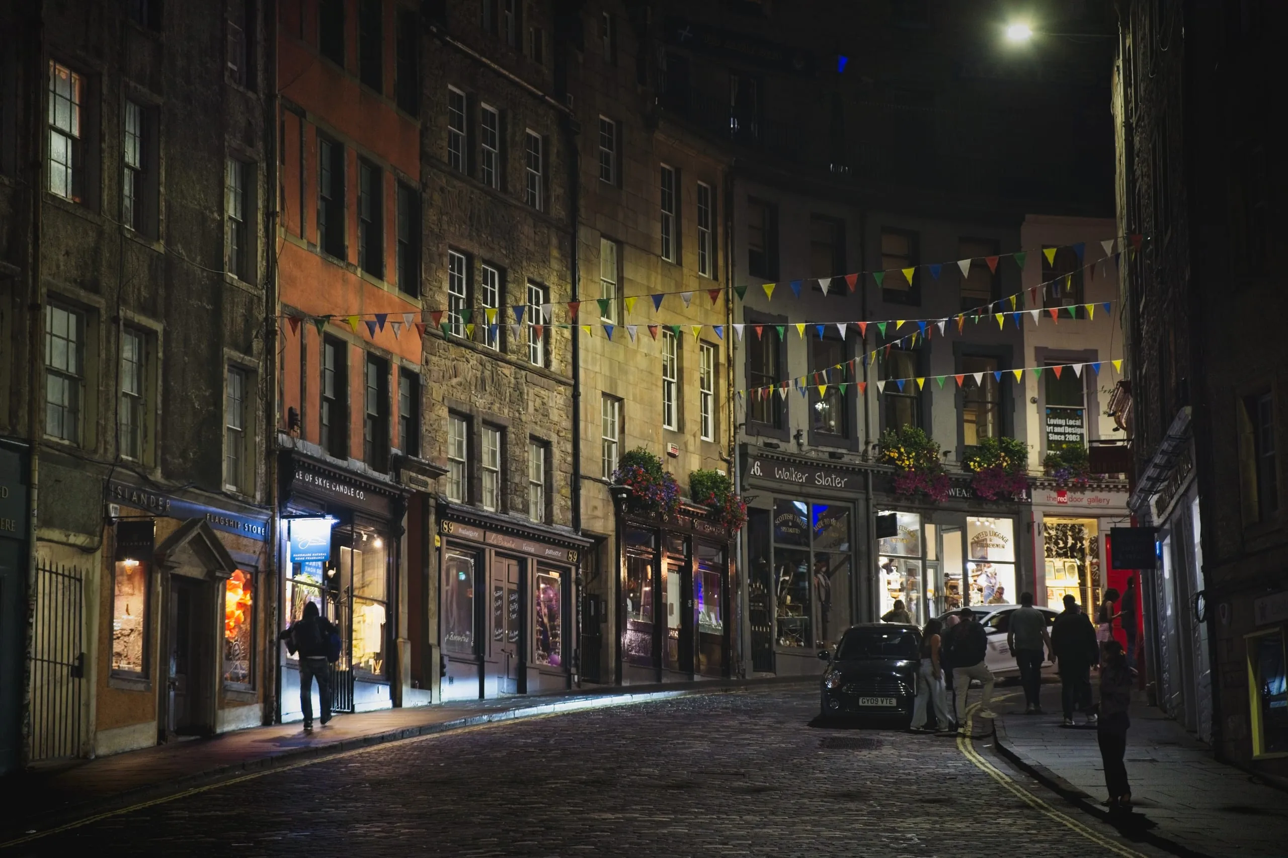 A color photograph of a cobblestone street in Edinburgh's Old Town at night, warmly lit shopfronts and bunting overhead as pedestrians and a parked car occupy the quiet road.