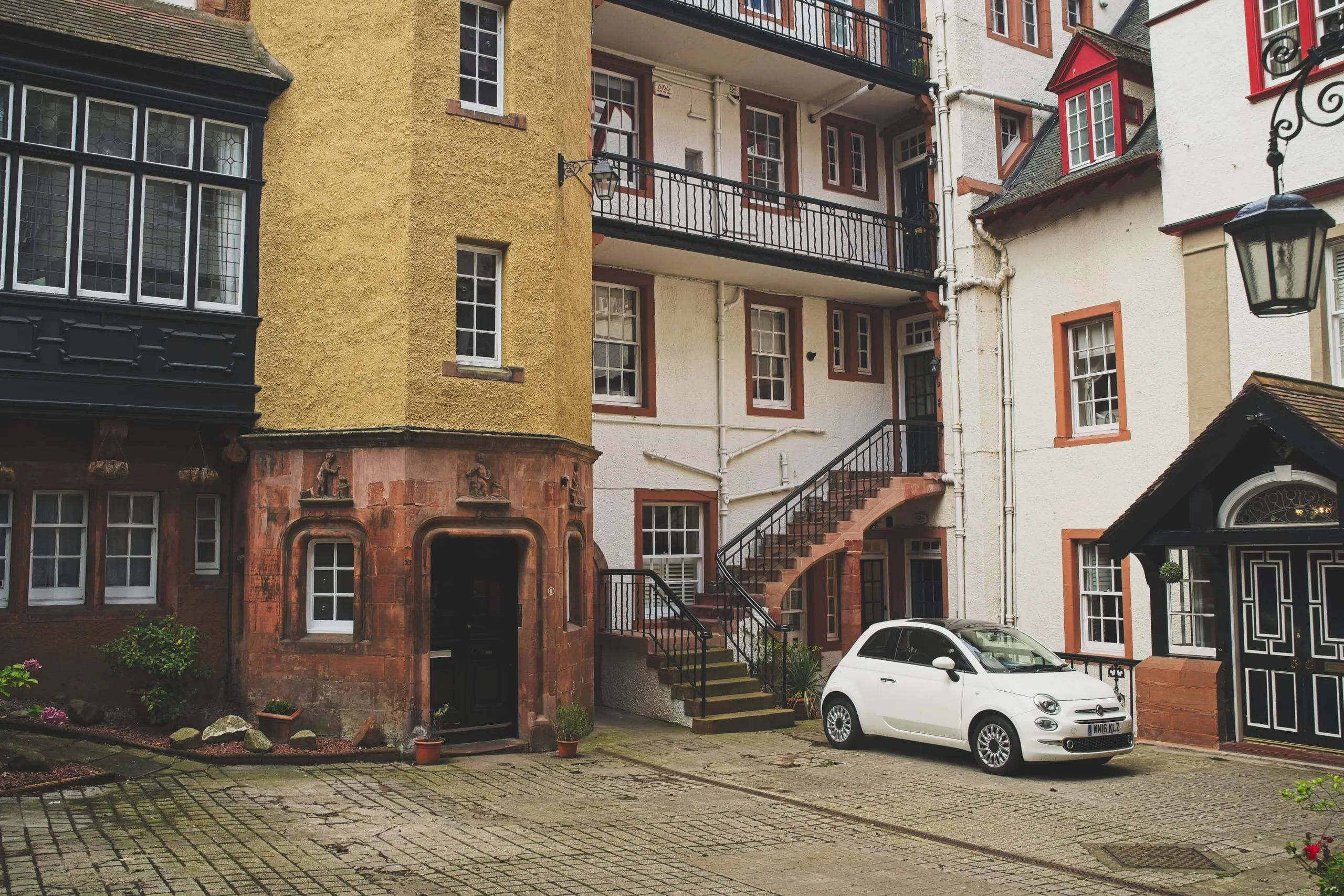 A color photograph of a stone courtyard in Edinburgh's Old Town by day, its ochre and white facades trimmed in red sandstone, with wrought-iron balconies, a curved staircase, and a small white car parked beside the doorway.