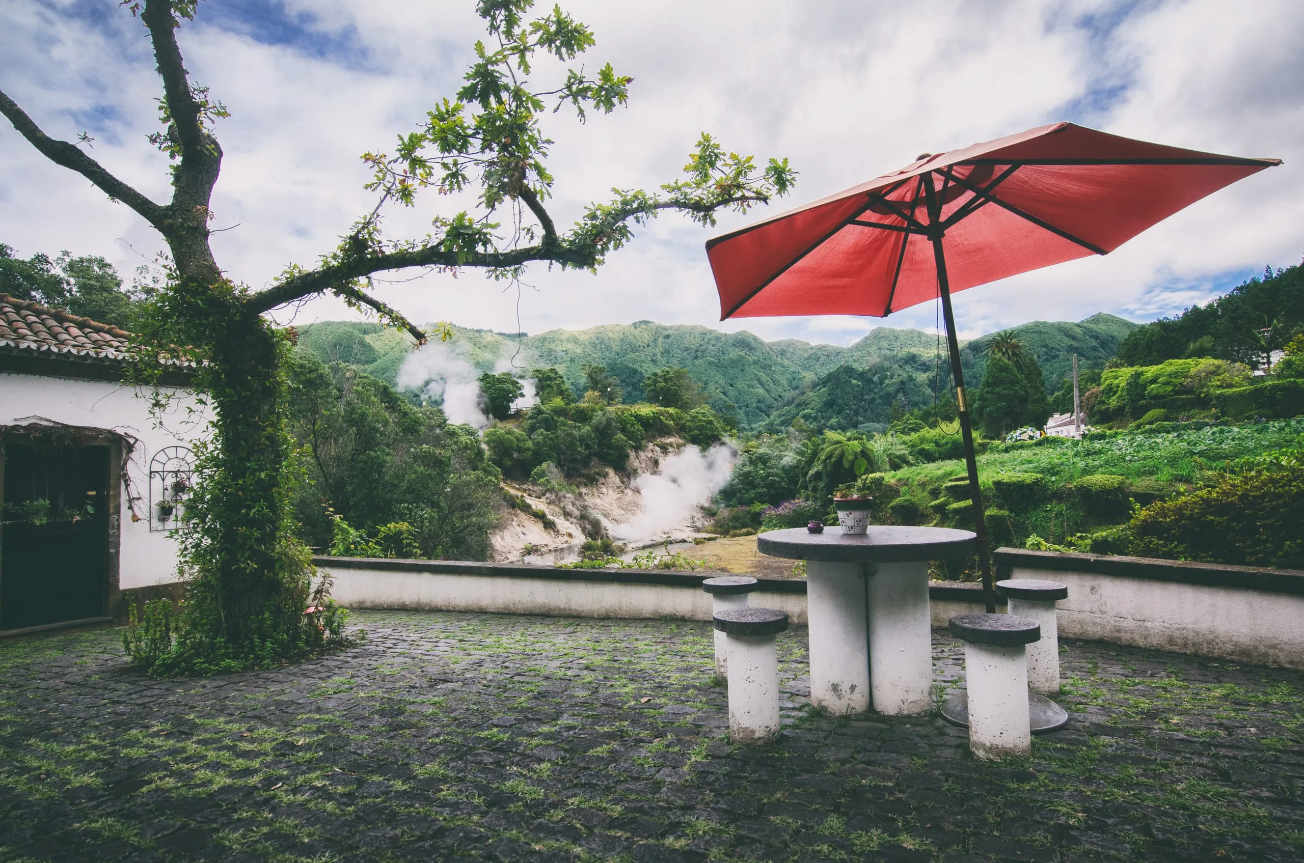 A red umbrella shades a stone table and stools on a cobbled terrace, with geothermal steam rising from a hillside beyond.