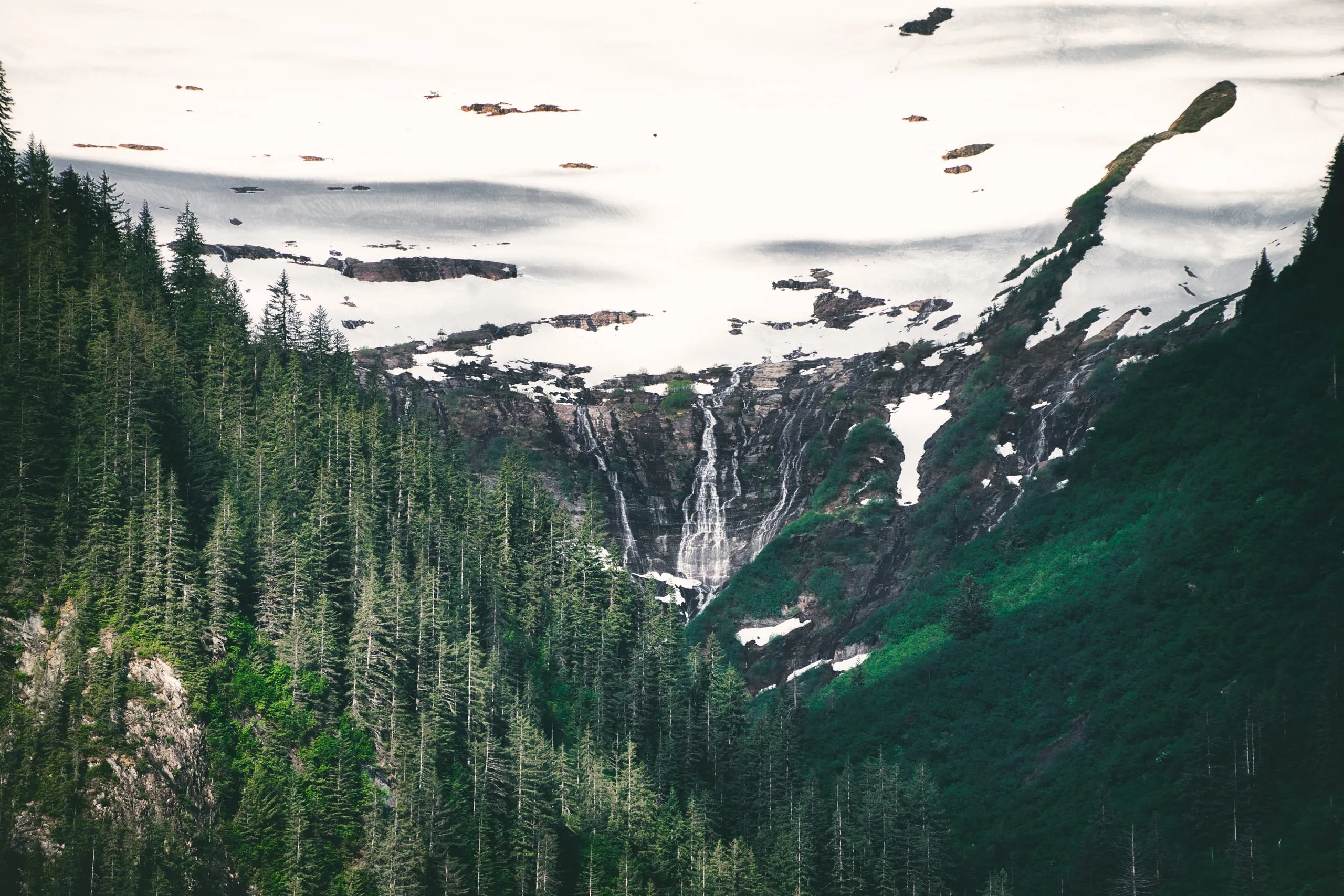 A braided waterfall spills from a broad snowfield down a dark cliff into a dense spruce forest below.