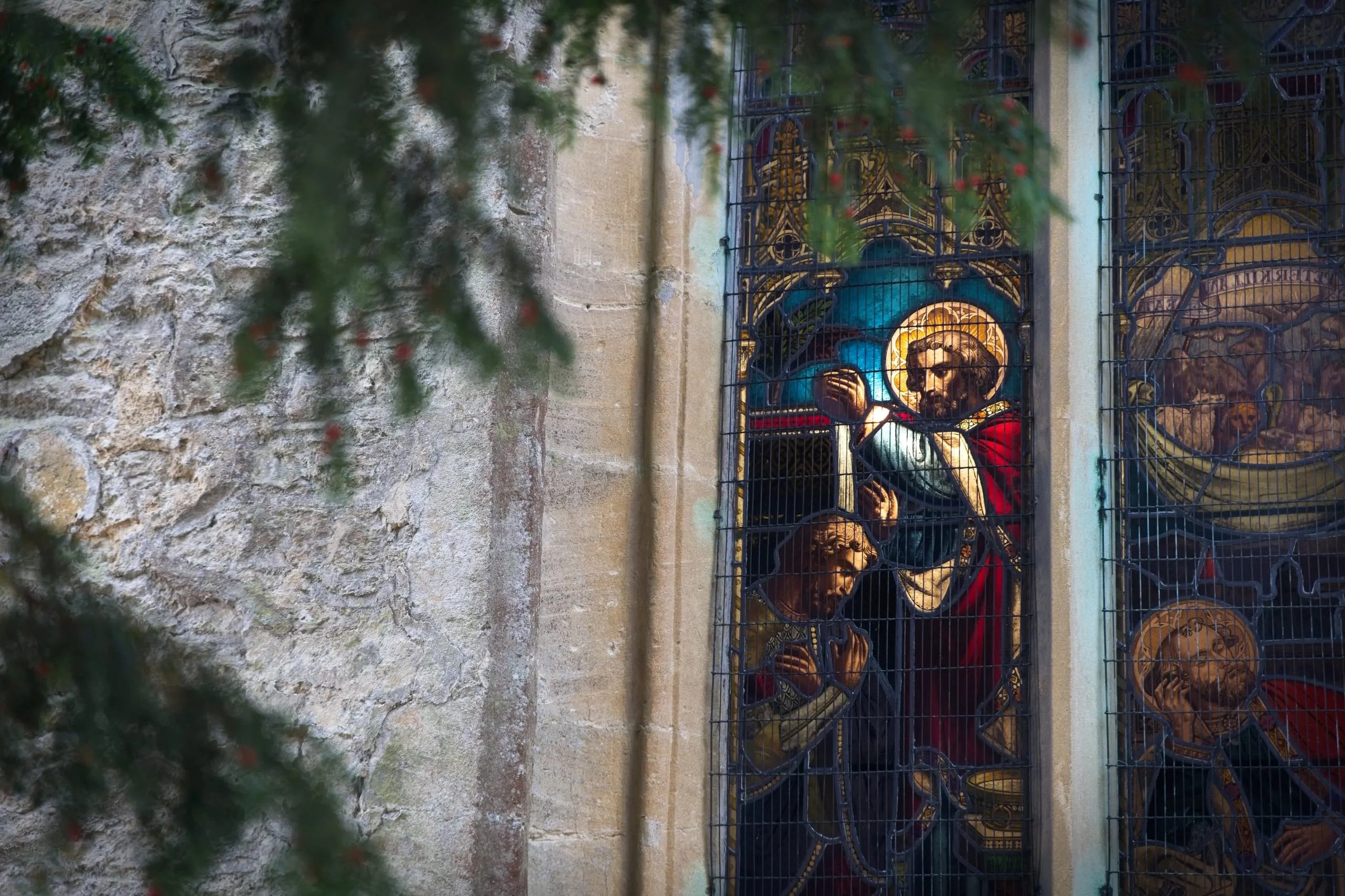 A photograph of a stained-glass window depicting a haloed saint blessing a kneeling figure, on the stone exterior of St Edward's Church in Stow-on-the-Wold, the Cotswolds, England, framed by yew branches.