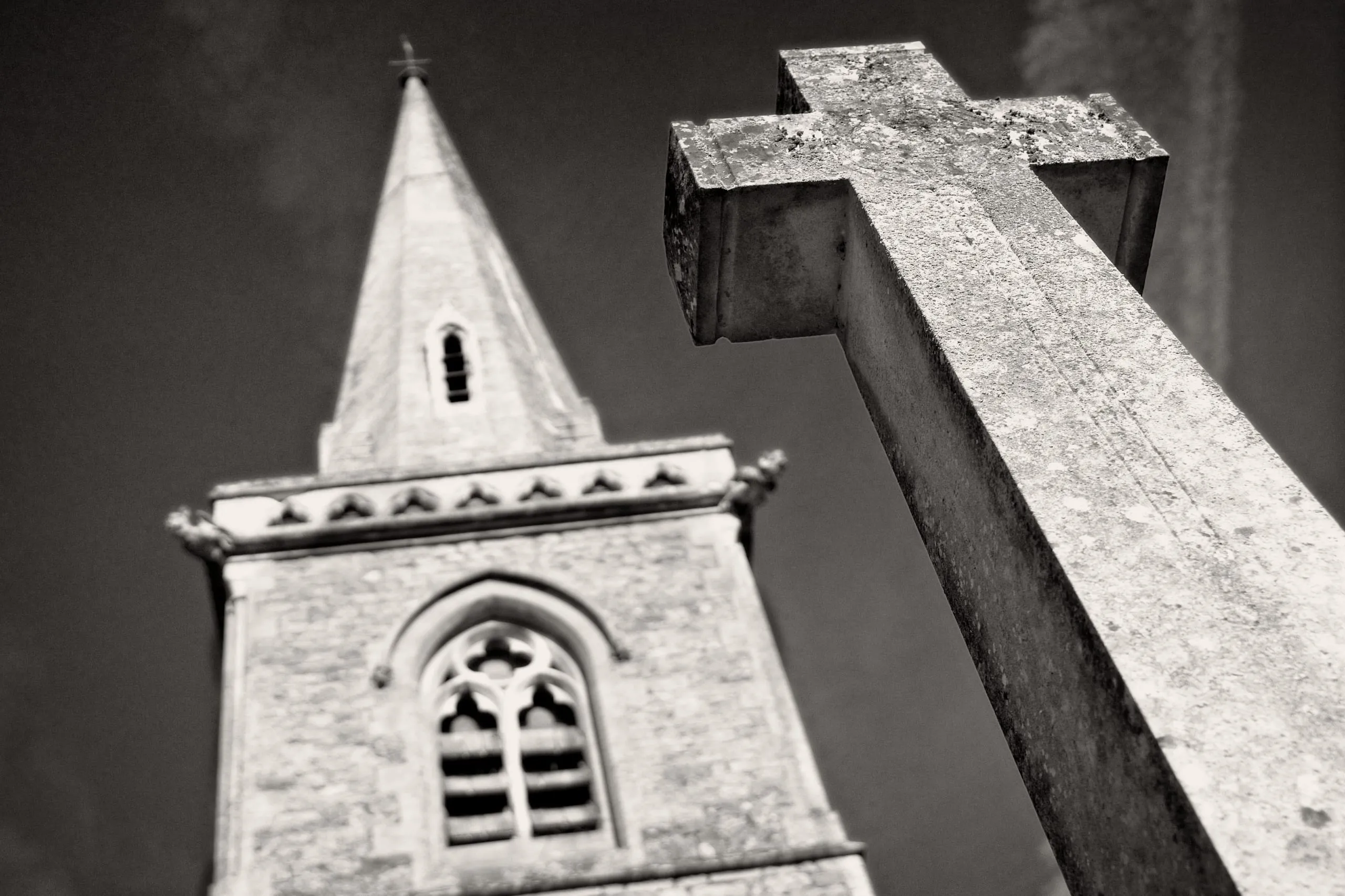 Black and white photograph of a weathered stone cross in sharp focus with a church steeple blurred in the background, in the Cotswolds, England.
