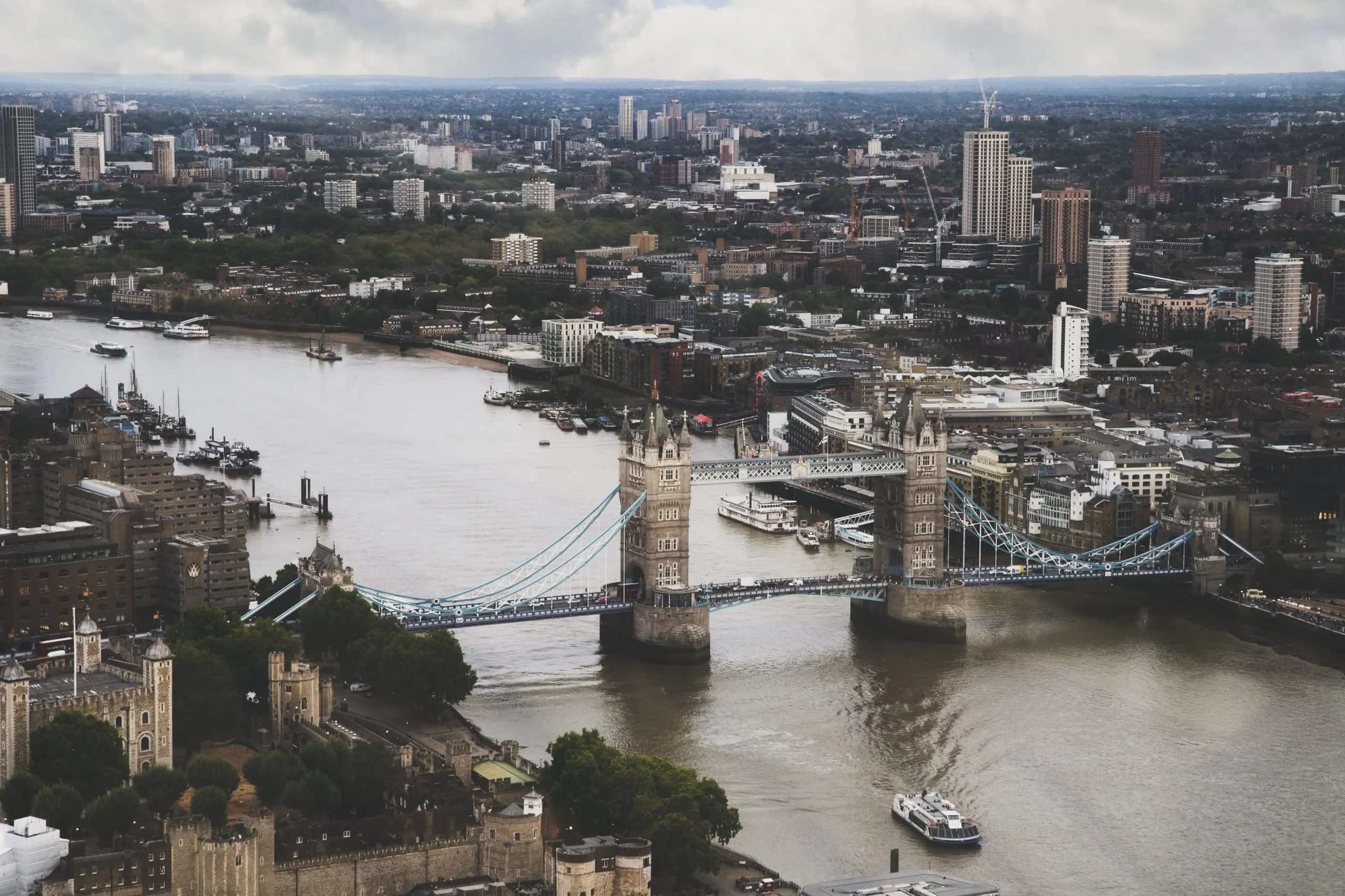 An aerial photograph of Tower Bridge spanning the River Thames in London, with the Tower of London in the foreground and boats moving along the water.