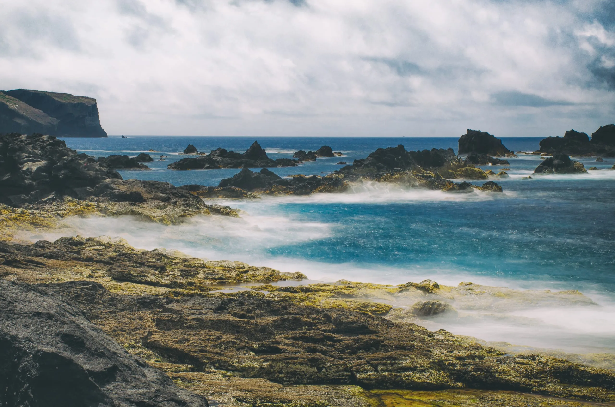 Long-exposure surf washing milky over dark volcanic rocks into a turquoise pool, with a headland cliff under cloudy sky.