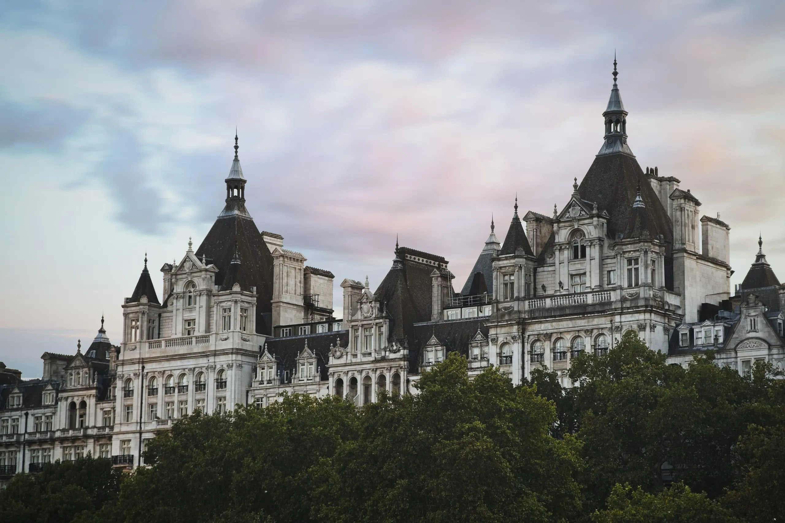 Photograph of the ornate turrets and mansard roofs of Whitehall Court rising above trees along the Thames under a pastel dusk sky, London, England.