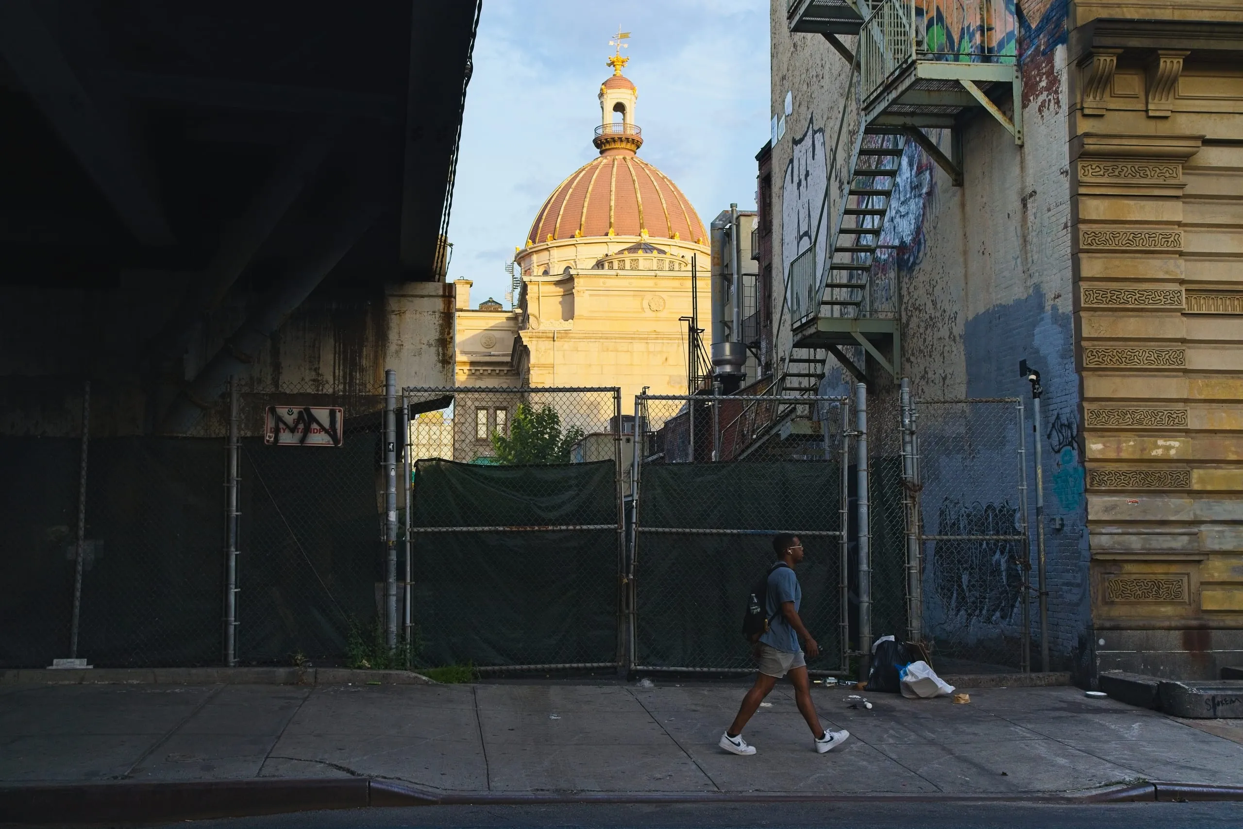 A color photograph of a man walking past a chain-link fence beneath elevated train tracks in Brooklyn, with the golden dome of a church lit by late sunlight in the background.
